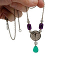 A necklace with a bronze coin shaped centerpiece etched with Sylvia Pankhurst's Angel of Freedom and Christabel Pankhurst's words, 'Take Courage,' with purple and green acrylic jewels in the style of the original Suffragette jewellery.