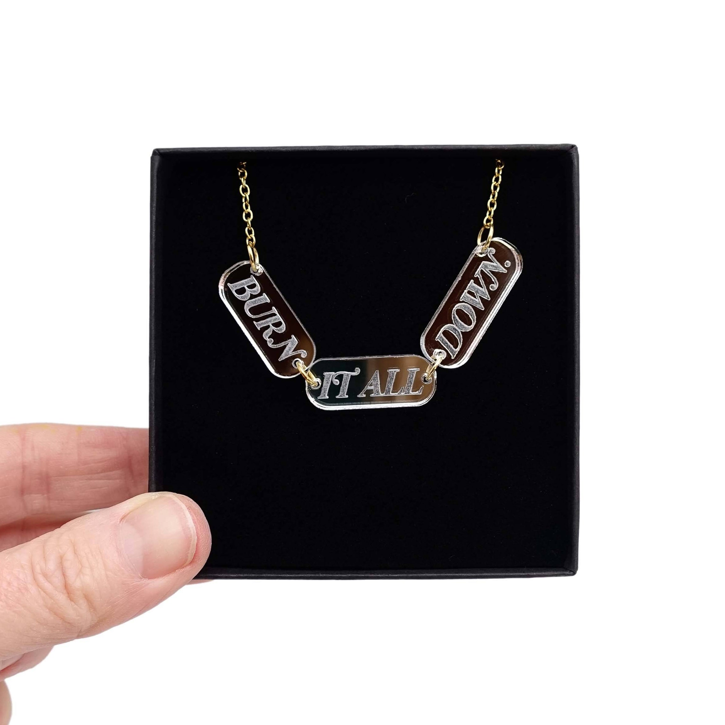 The BURN IT ALL DOWN necklace in silver mirror on a gold-plated stainless steel chain, shown held up in a Wear and Resist gift box.