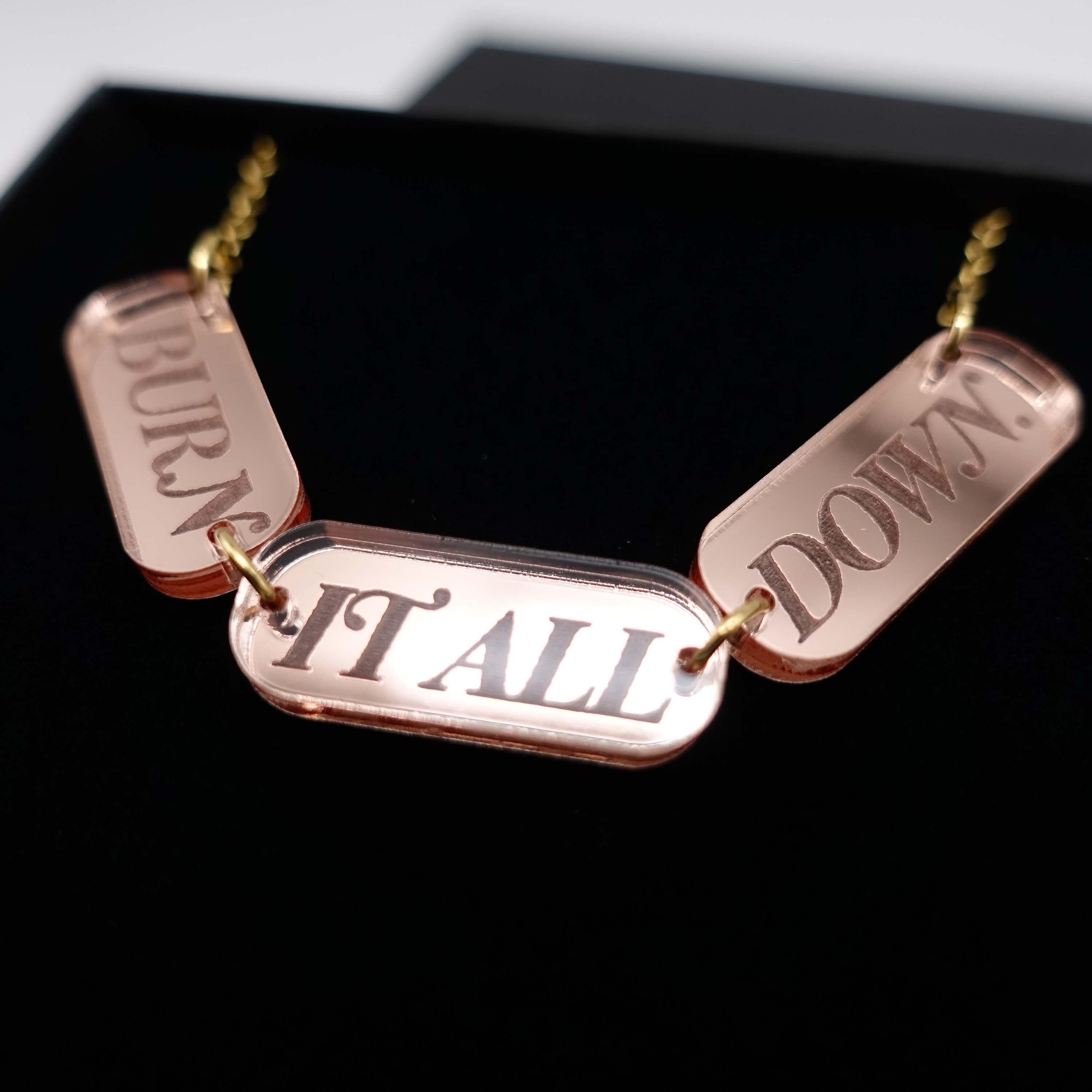 A close up of the BURN IT ALL DOWN necklace in rose-gold mirror on a gold-plated stainless steel chain, shown in a Wear and Resist gift box.