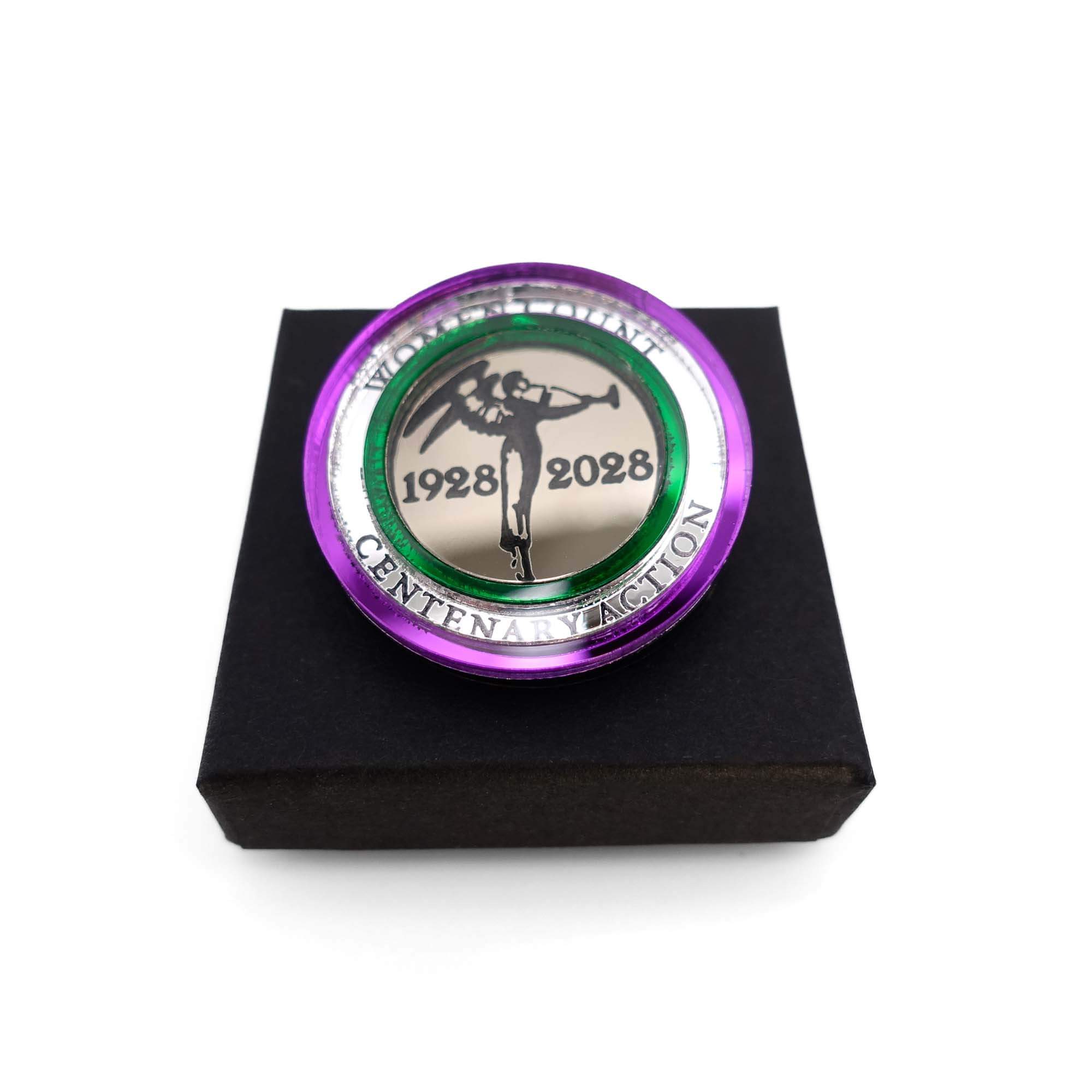 A brooch with a central medallion featuring Sylvia Pankhurst's angel of freedom, encircled by bands in the Suffragette colours and text including '1928-2028' and 'Women Count', 'Centenary Action' shown on a Wear and Resist gift box.