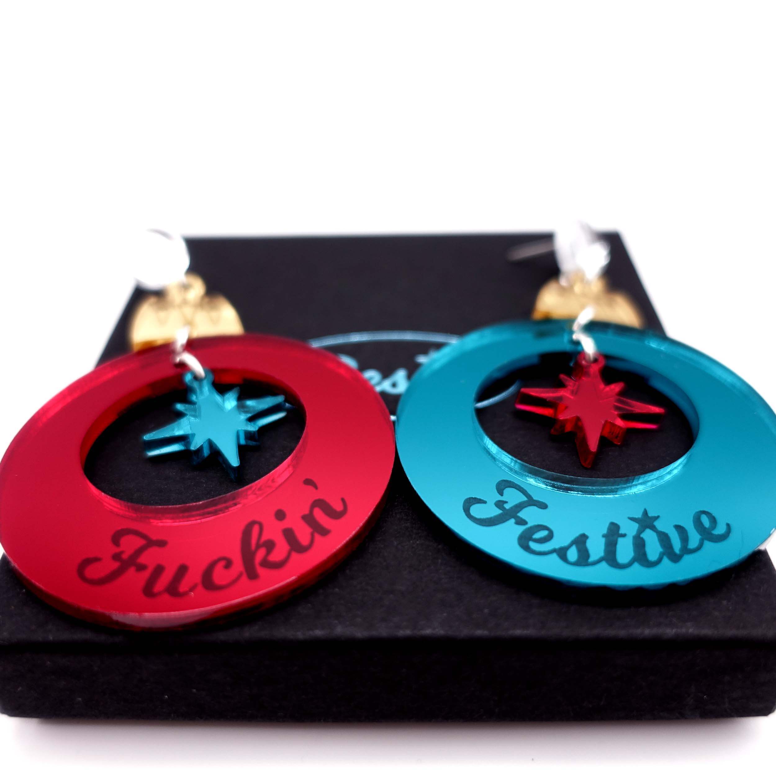 A close up of the F*cking Festive Christmas bauble earrings in ruby and teal mirror, shown on a Wear and Resist gift box.