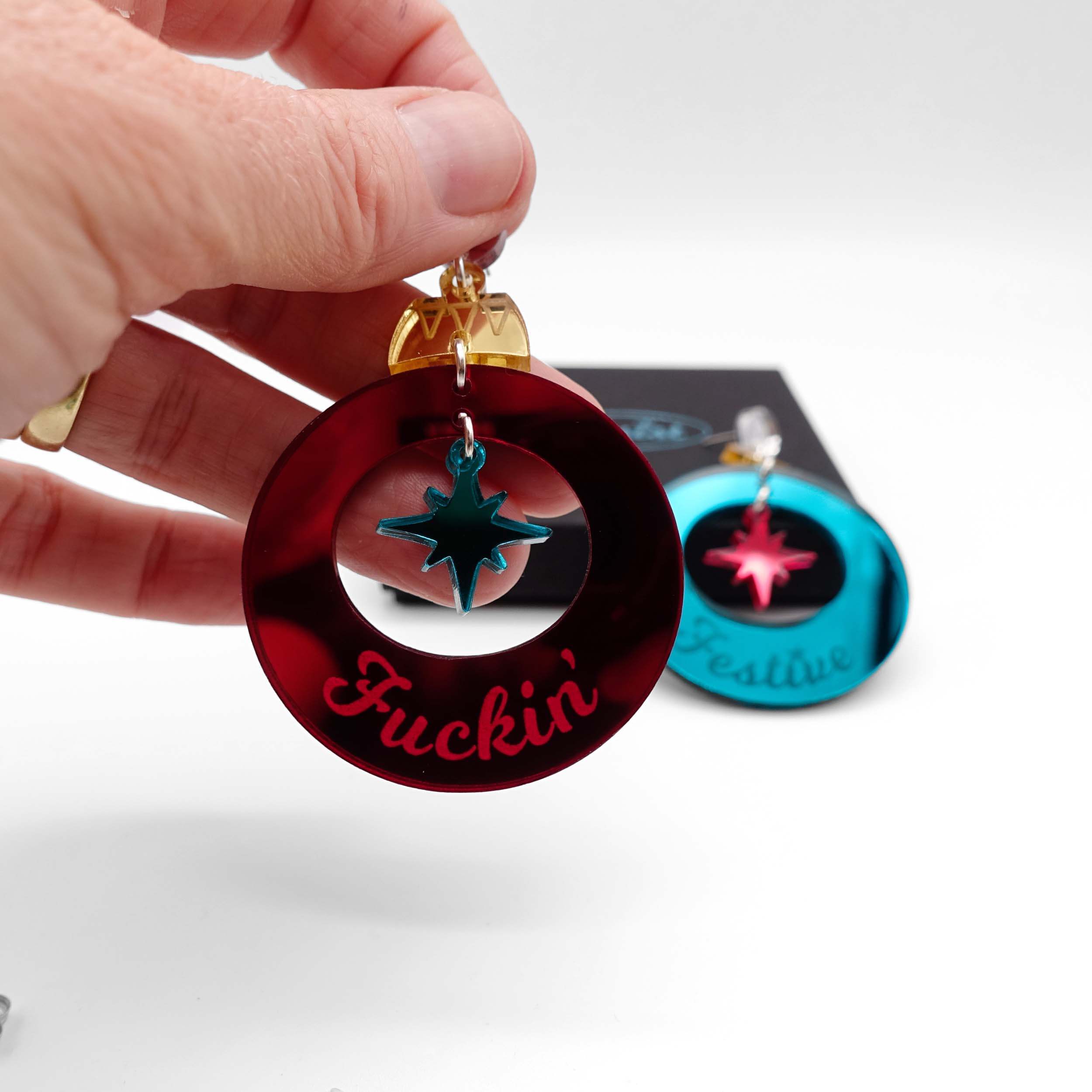 F*ckin Festive Christmas bauble earrings in ruby and teal mirror, with the ruby F*ckin shown held up in front of the teal Festive resting on a Wear and Resist gift box.