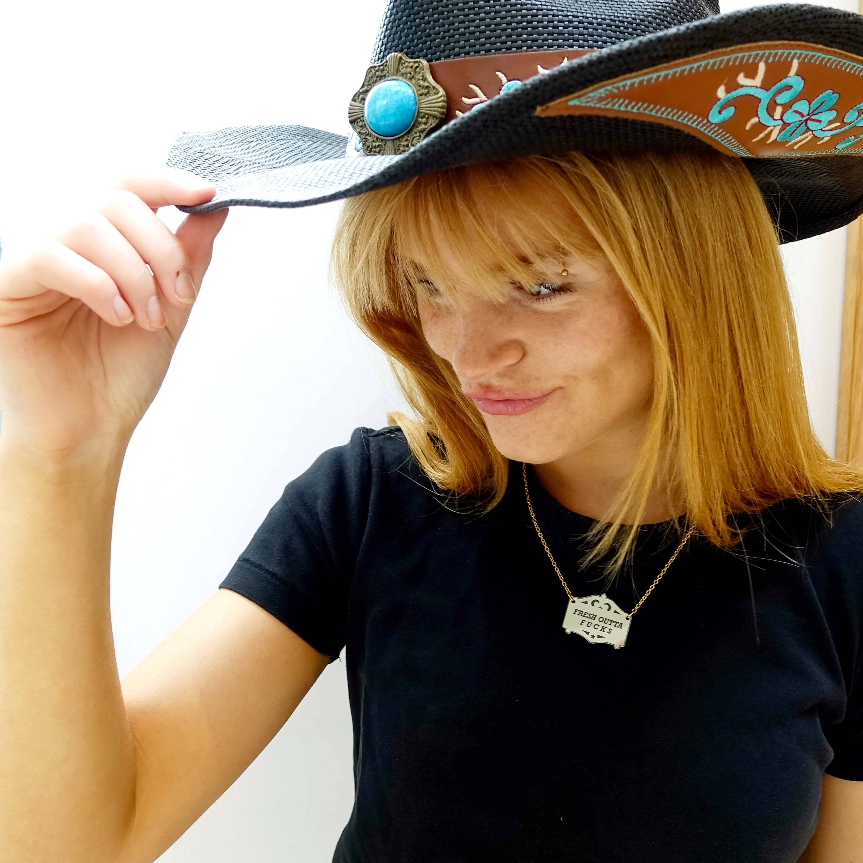 Eliza tips her cowboy hat, wearing a bronze Fresh outta f*cks mirror cowboy necklace on a gold-plated stainless steel chain.