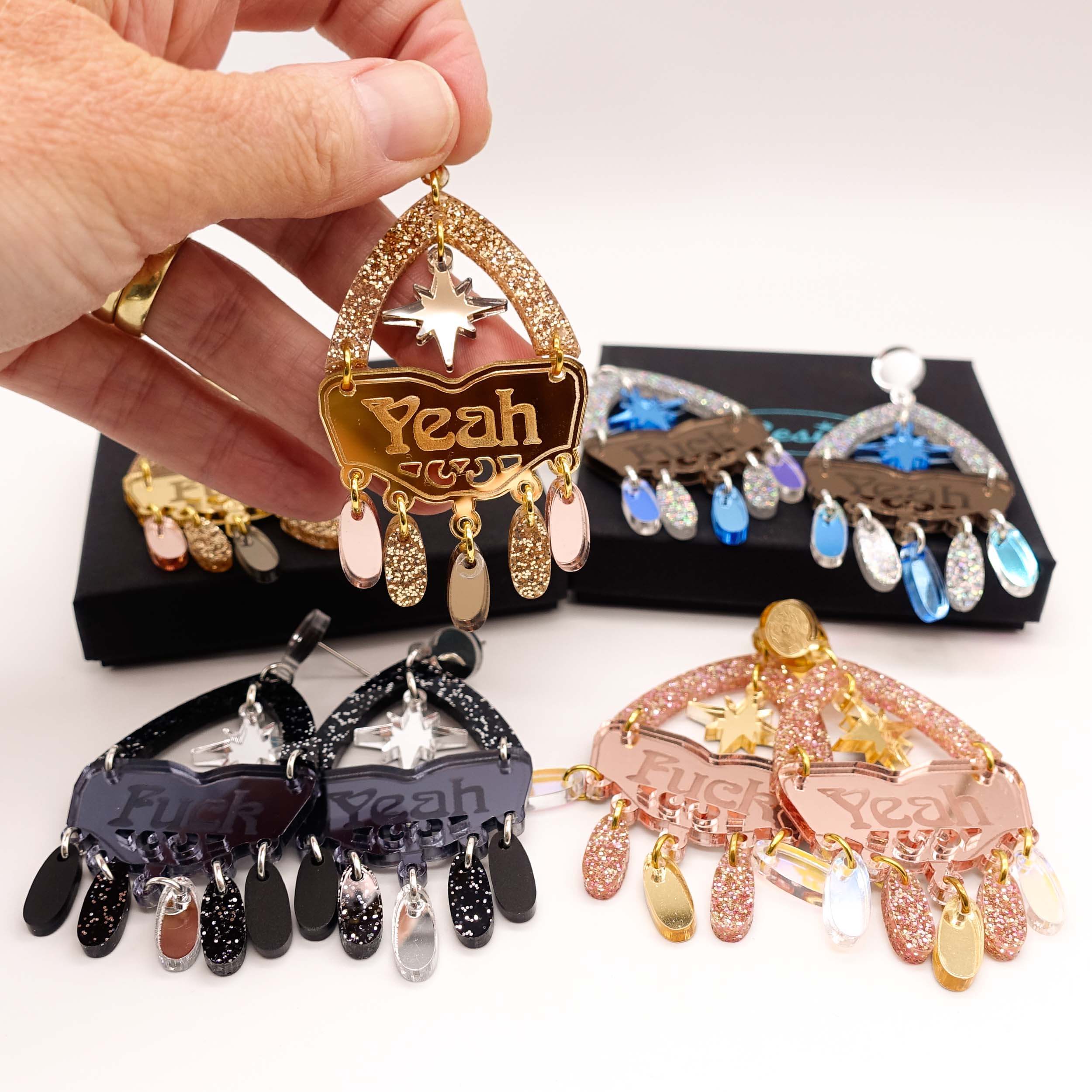 All four colourways of the F*ck Yeah statement earrings with my hand holding up one earring for scale.
