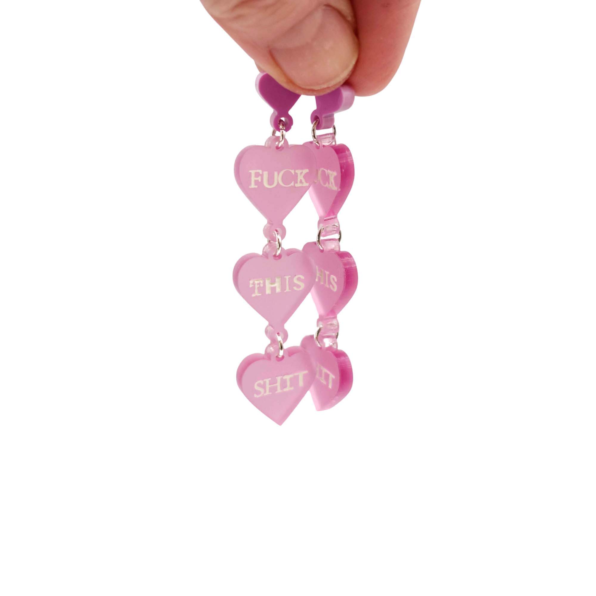 F*ck this Sh*t earrings in pale pink with glow-in-the-dark words. Shown held up against a white background.