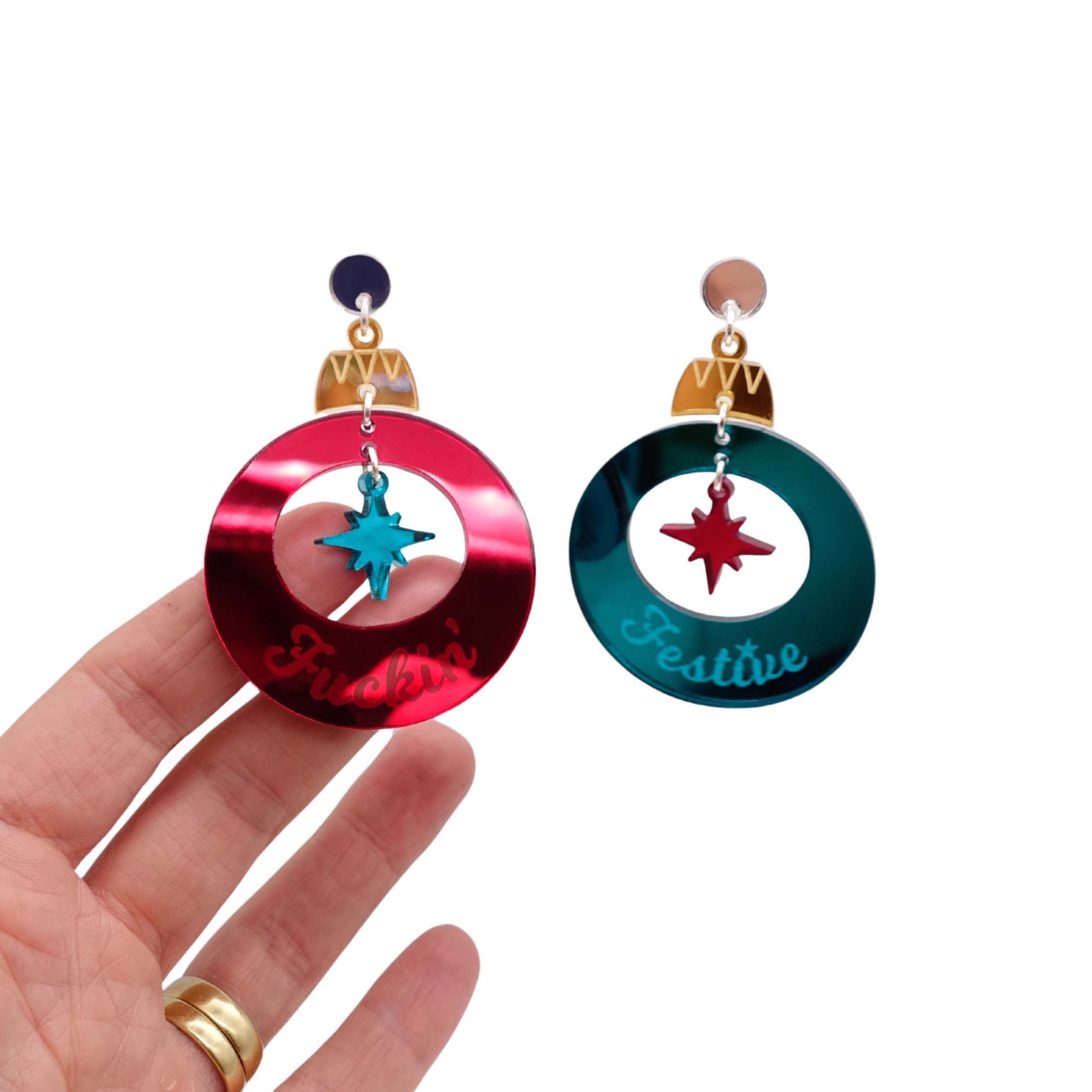 F*ckin Festive Christmas bauble earrings in ruby and teal mirror, shown hanging against a white background, with my hand behind for scale and to show the reflective nature of the mirror acrylic. Happy sweary Christmas!