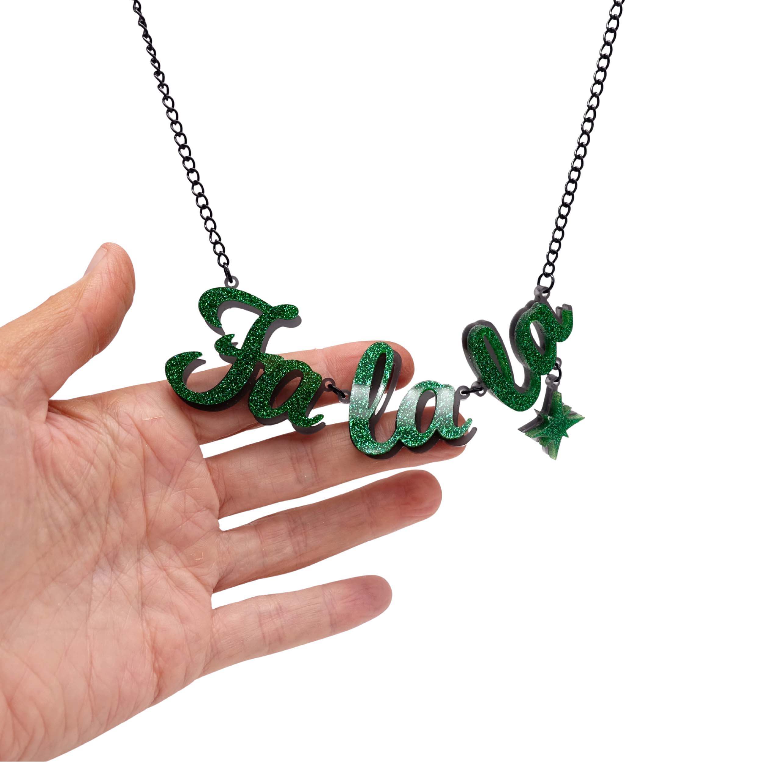 Fa La LA necklace in green glitter, shown hanging against a white background with my hand behind for scale.