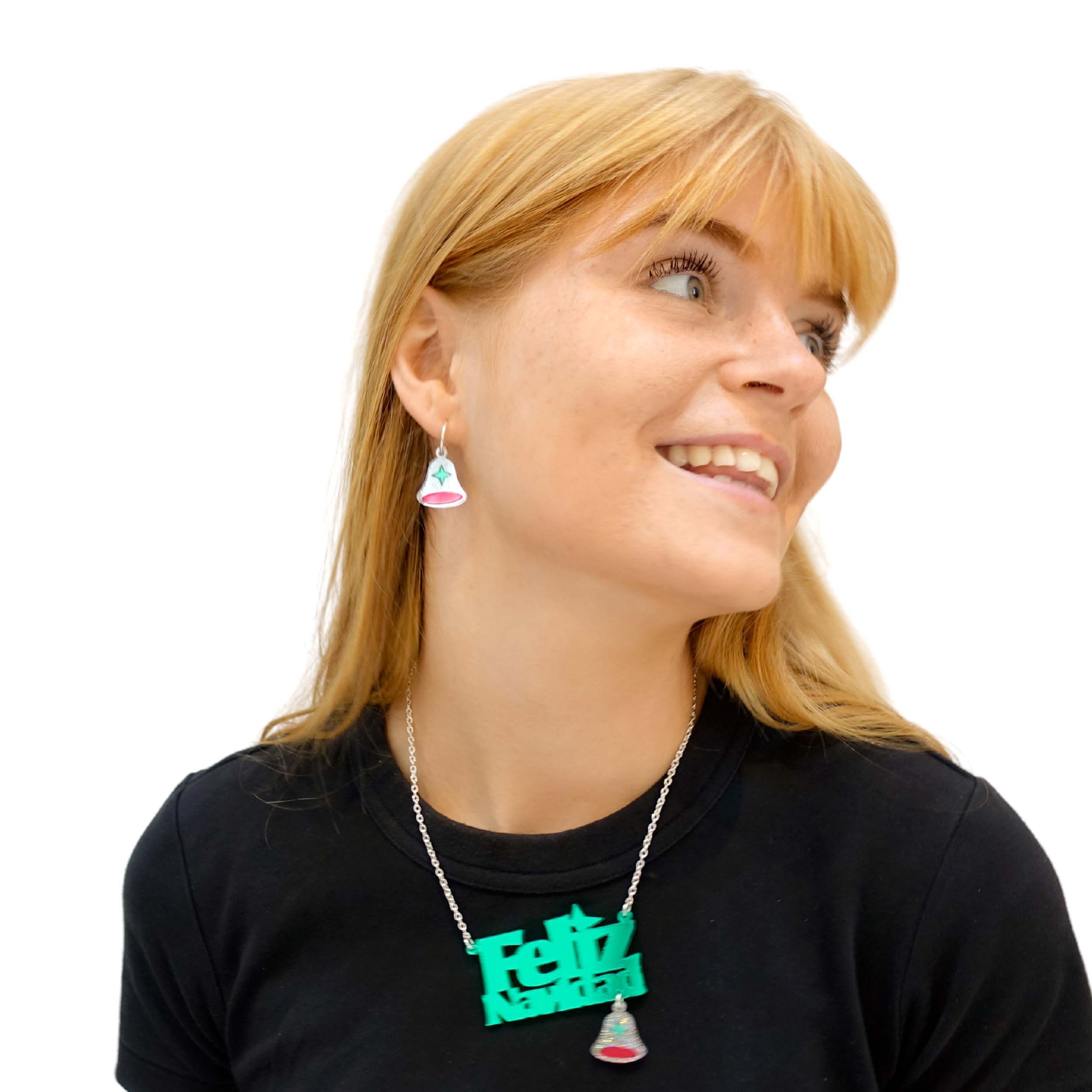 Eliza wears silver glitter bell hoop earrings and a Feliz Navidad necklace with hanging silver glitter bell.