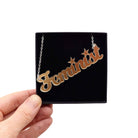 Script font Feminist necklace in matte iridescent, held up in a Wear and Resist gift box. A great gift for a fellow feminist!