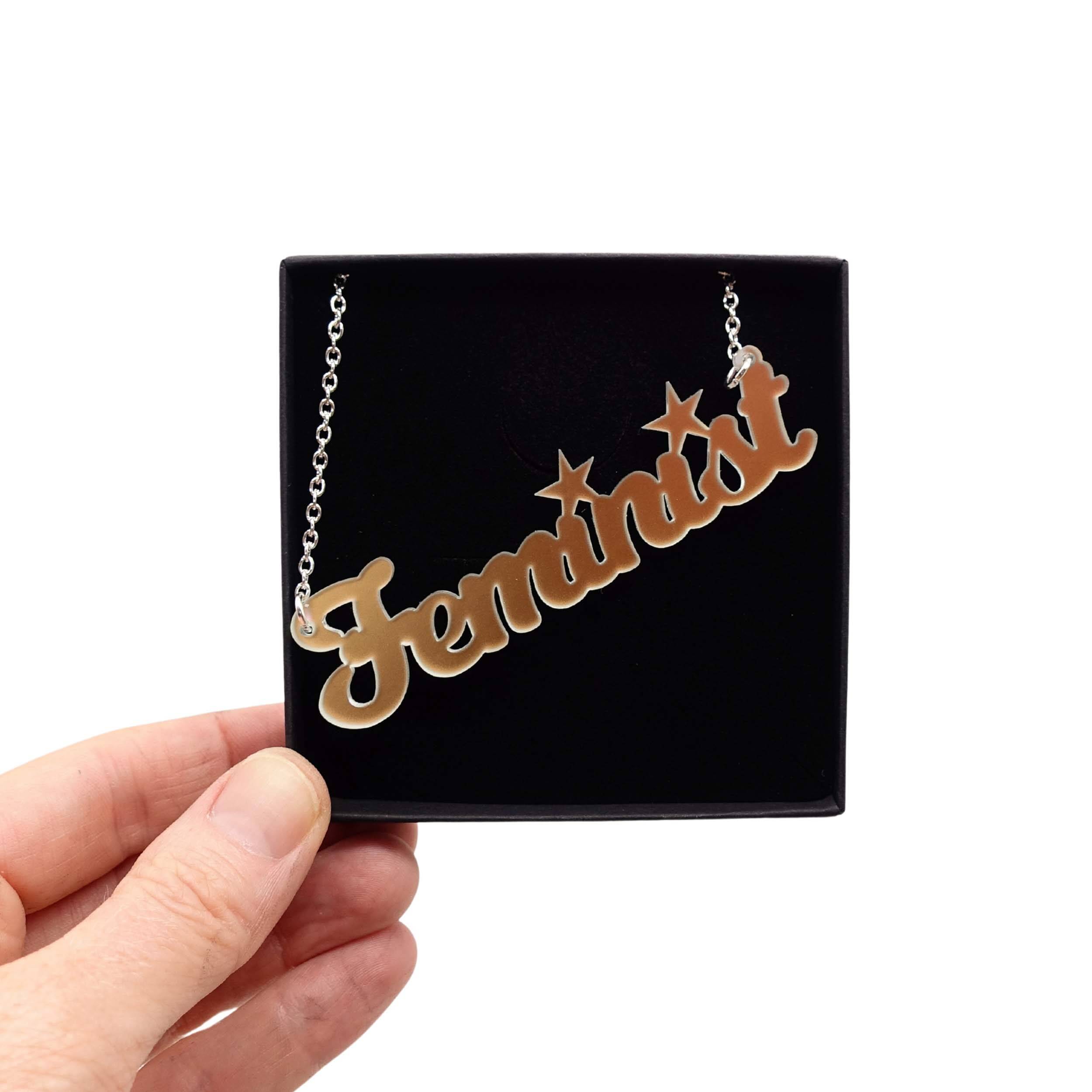 Script font Feminist necklace in matte iridescent, held up in a Wear and Resist gift box. A great gift for a fellow feminist!