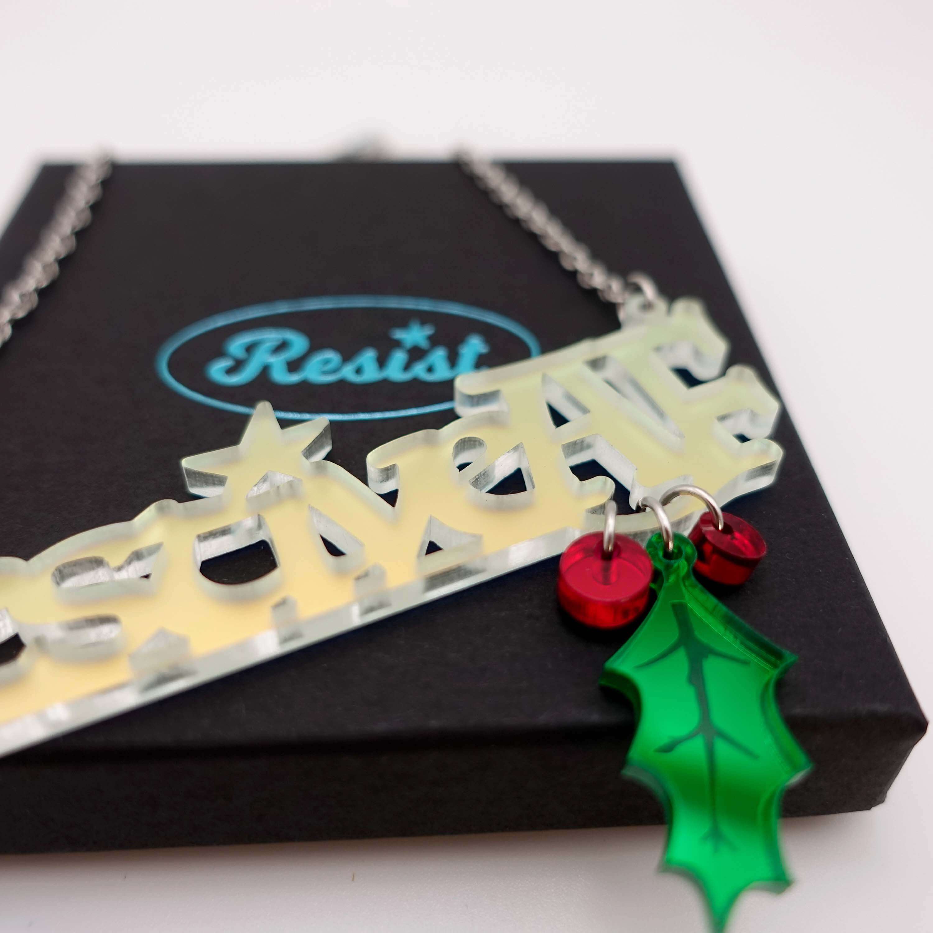 A close up of the Festive AF matte iridescent necklace with hanging holly leaf and berries, shown sitting on a Wear and Resist gift box.