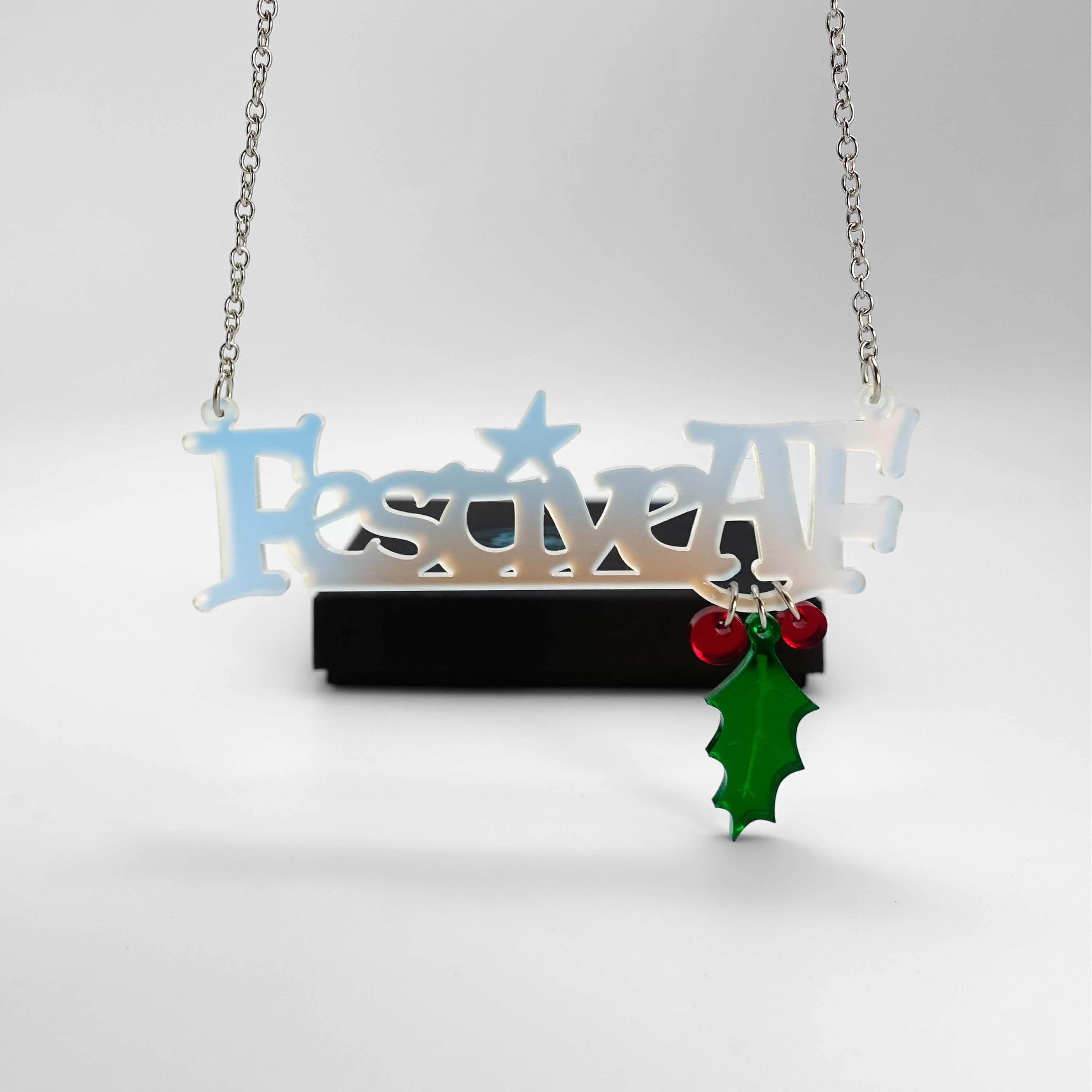 Festive AF matte iridescent necklace with hanging holly leaf and berries, shown hanging in front of a Wear and Resist gift box.