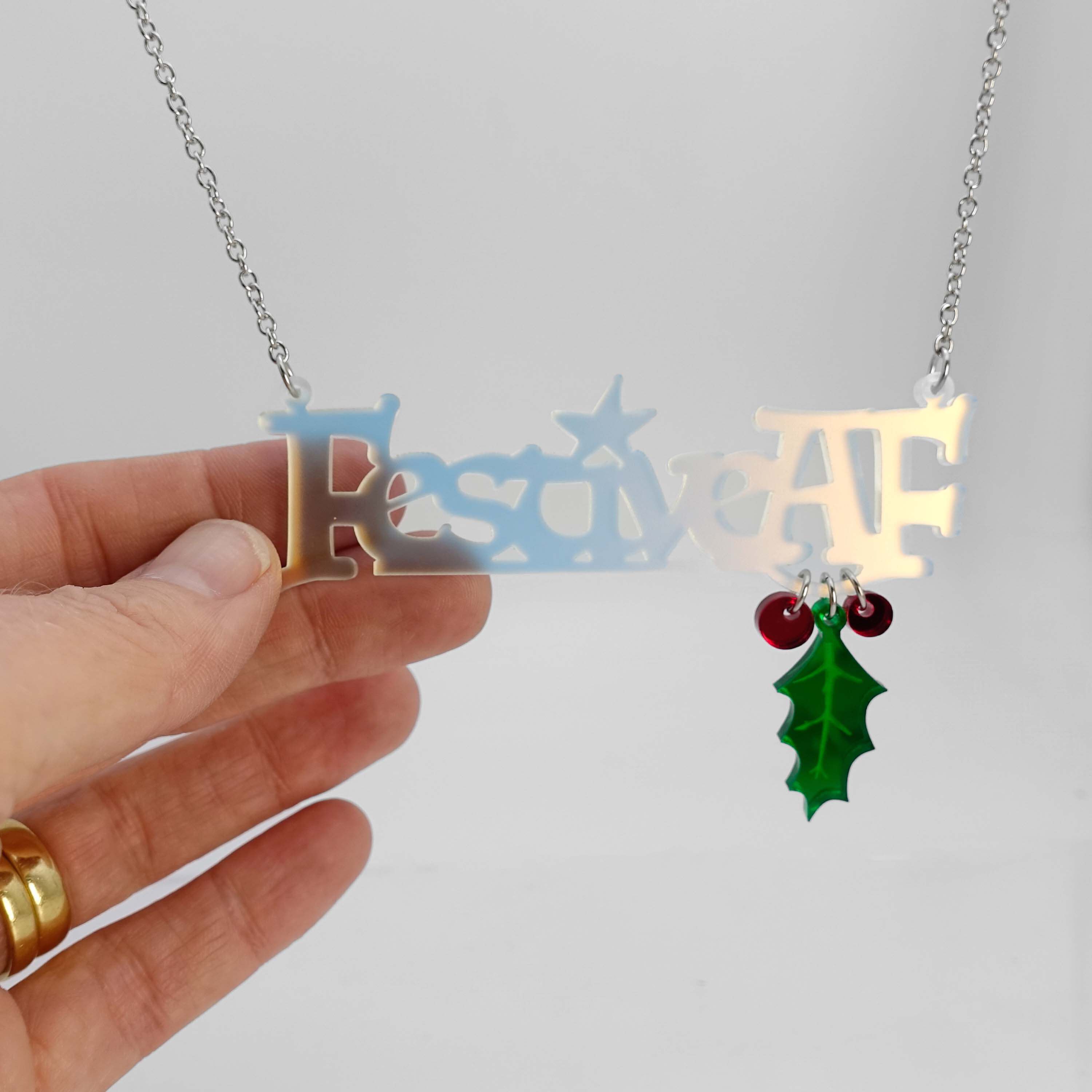 Festive AF matte iridescent necklace with hanging holly leaf and berries, shown hanging catching the light with my hand holding it for scale.