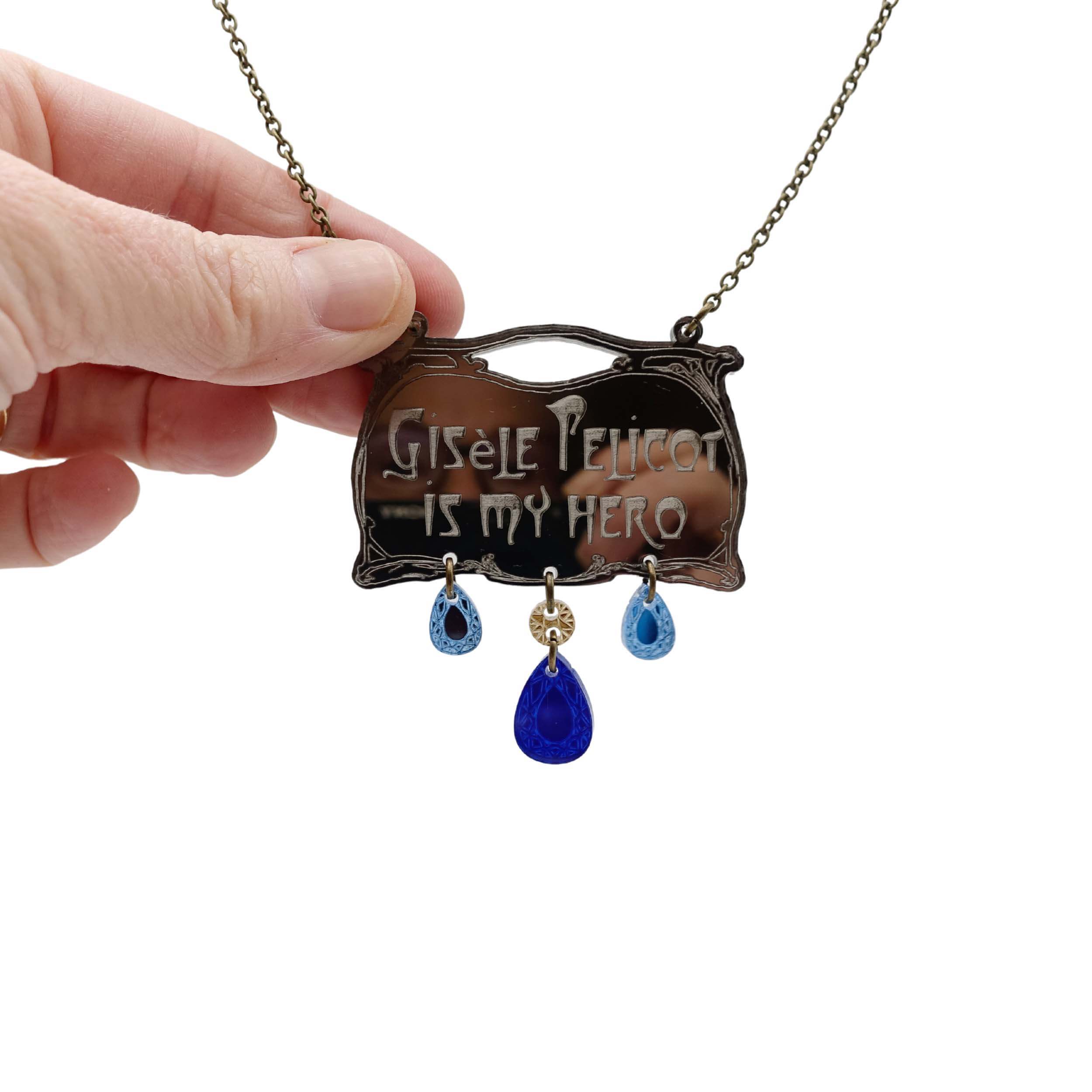Bronze necklace in Paris Metro Hector Guimard style with etched writing 'Gisele Pelicot is my hero' with dangling acrylic jewels in sky and electric blue shown with my hand foro scale hanging on an antique gold chain against a white background.