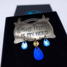 A close up of the Gisele Pelicot is My Hero necklace with hanging jewels shown in a Wear and Resist gift box.