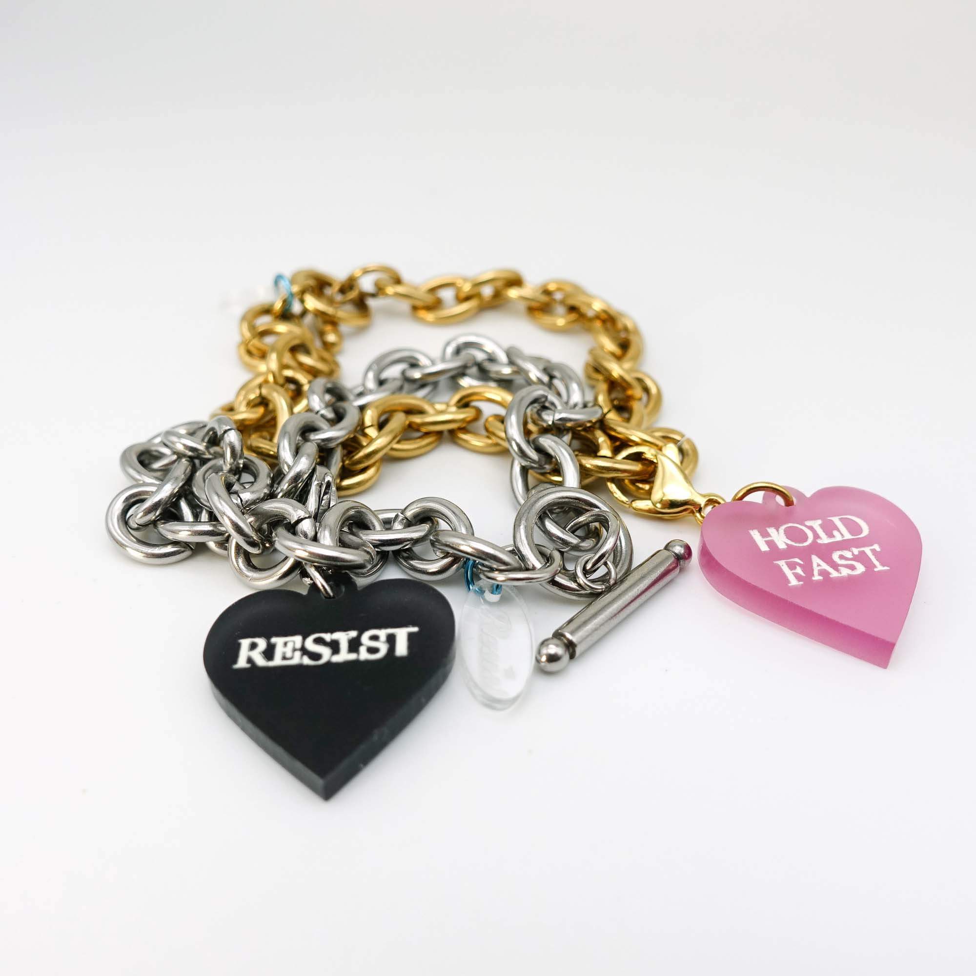 Charm bracelets shown on a white surface in stainless steel and gold with two charms etched in glow-in-the-dark pigment that say Resist and Hold Fast.