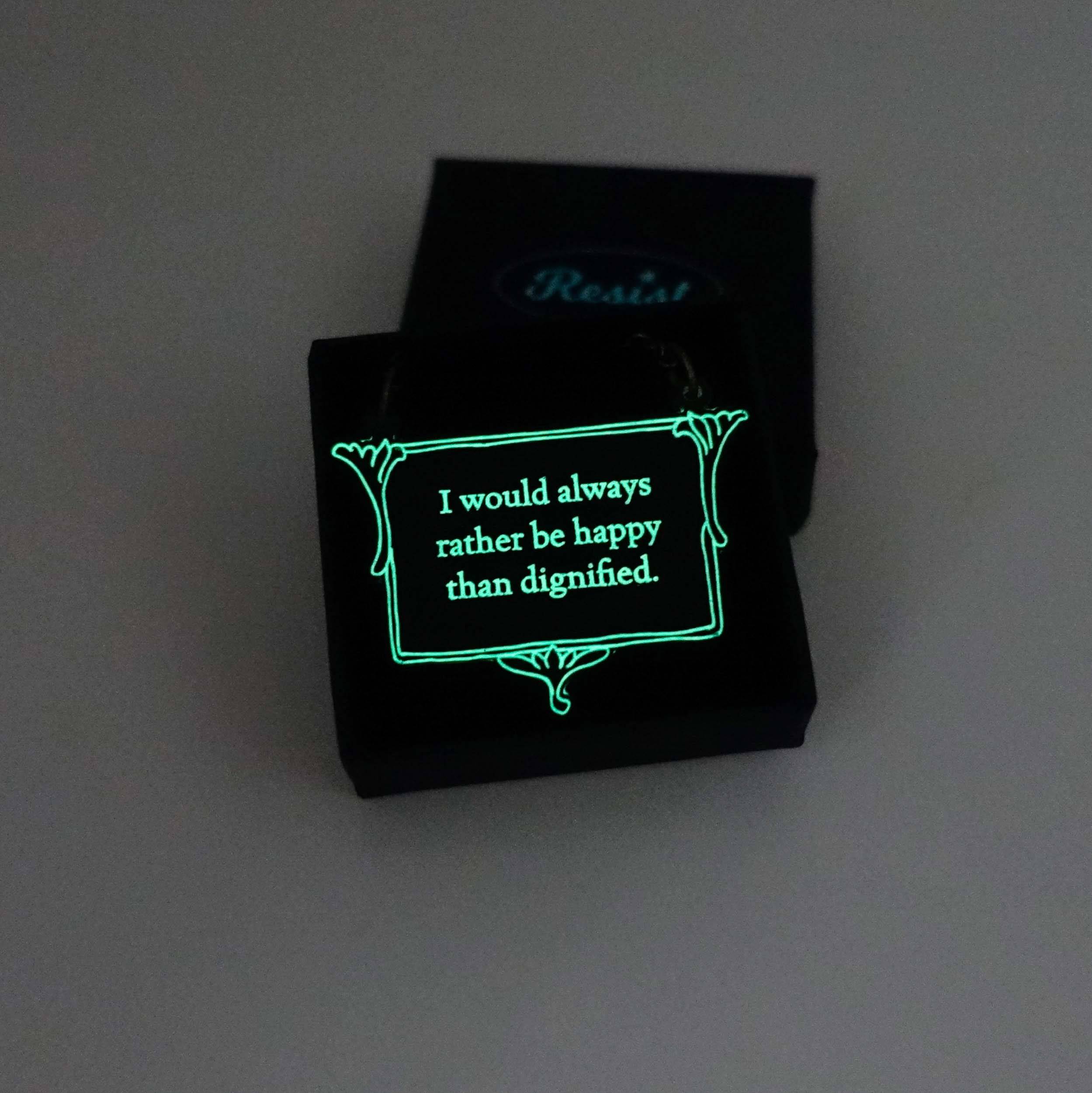 A vintage style slate necklace etched with the words of Jane Eyre 'I would always rather be happy than dignified', shown glowing in the dark in a small Wear and Resist gift box.