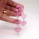 I Love Me heart drop earrings in pale pink frost with glow-in-the-dark etched words, shown held in my hand for scale.