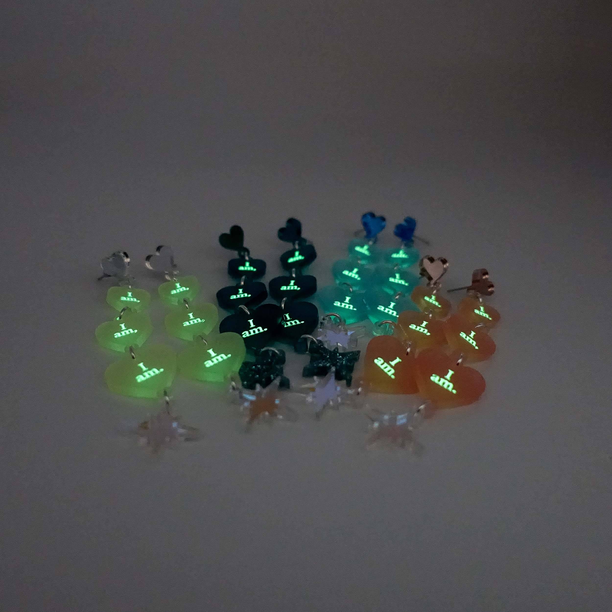 A group shot of all of the  I am I am I am heart drop earrings glowing in the dark.