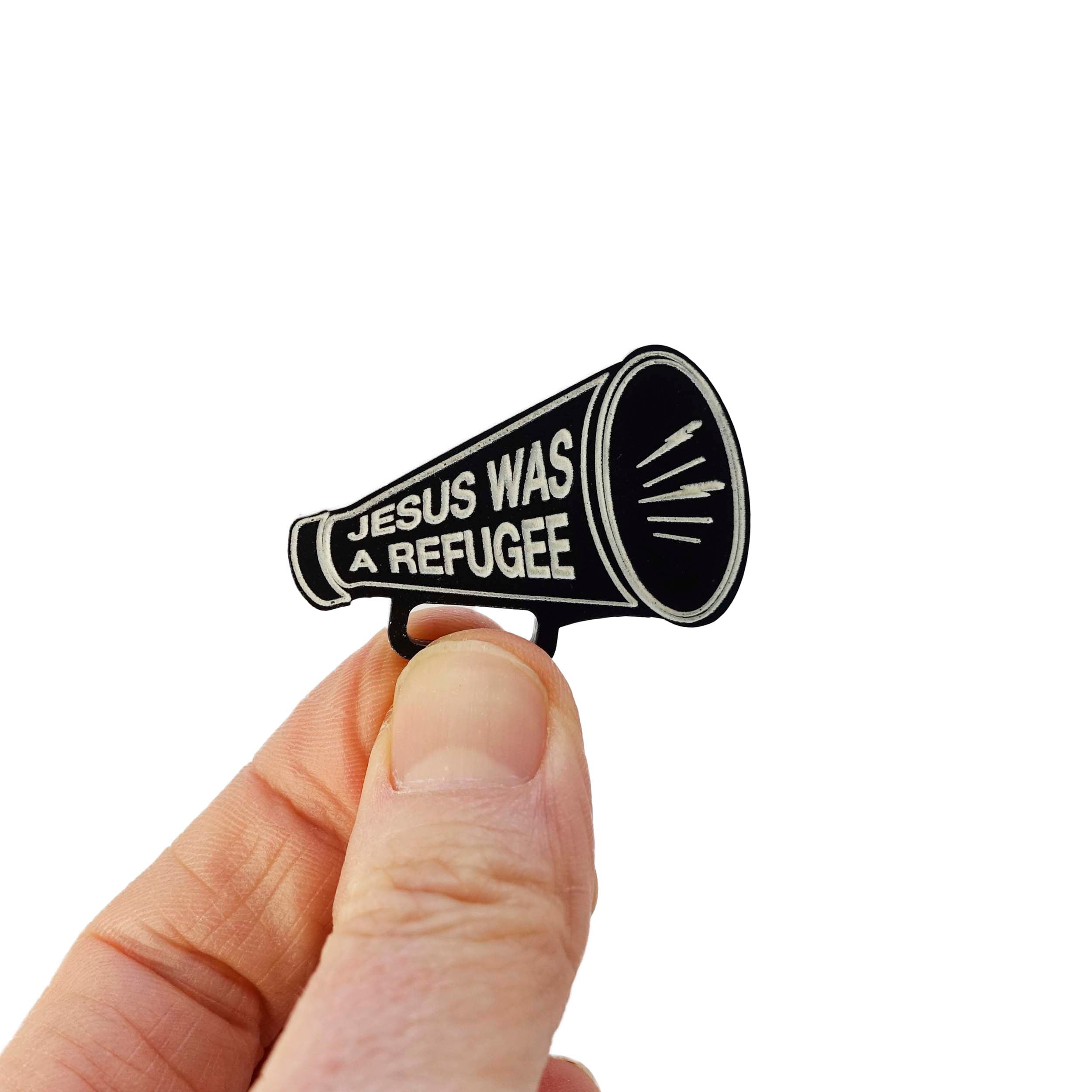 Black megaphone brooch etched in glow-in-the-dark paint with the words Jesus was a Refugee. Held up in my fingers for scale.