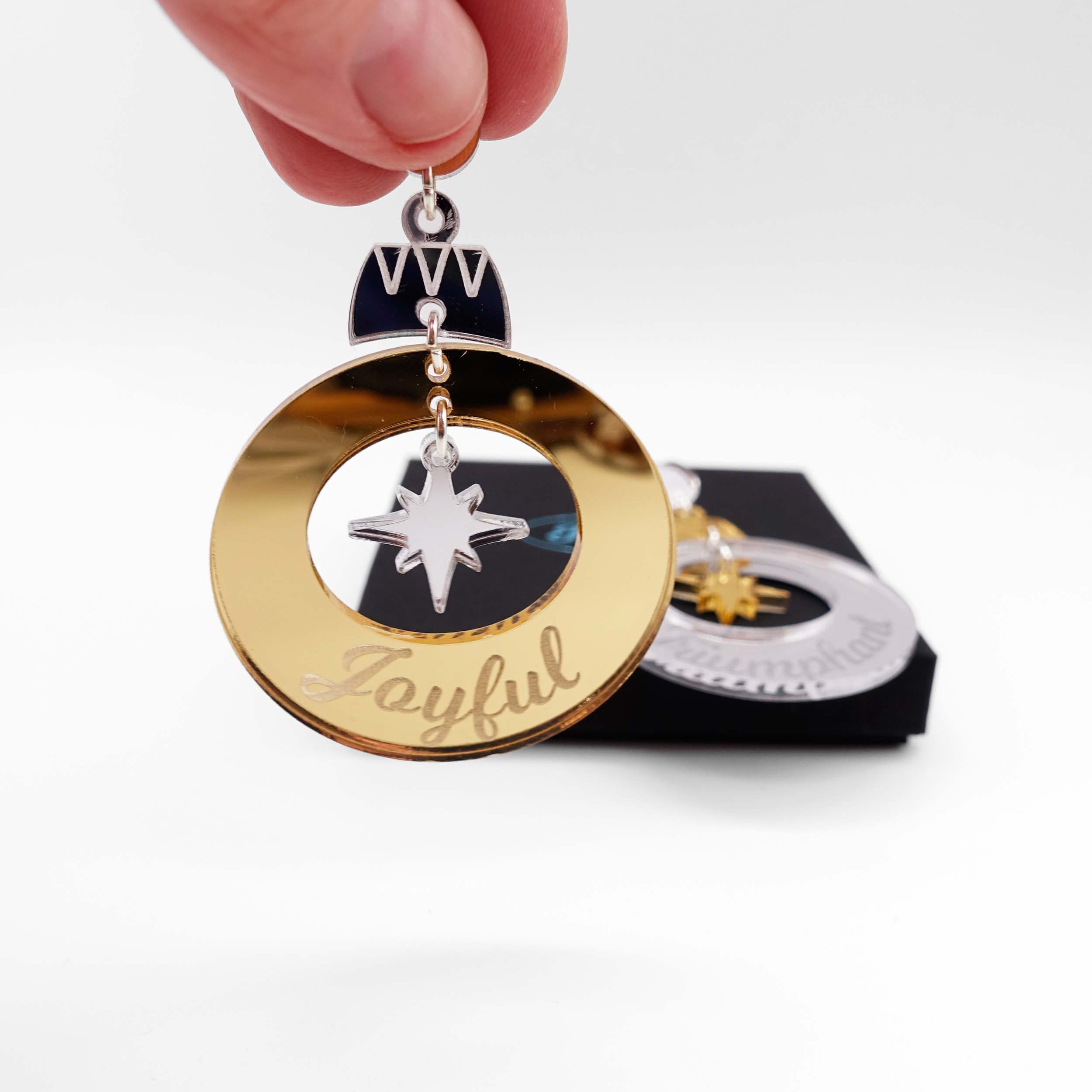 Joyful and Triumphant Christmas Bauble earrings in gold and silver, with one held up in front of the other, resting on a Wear and Resist gift box.