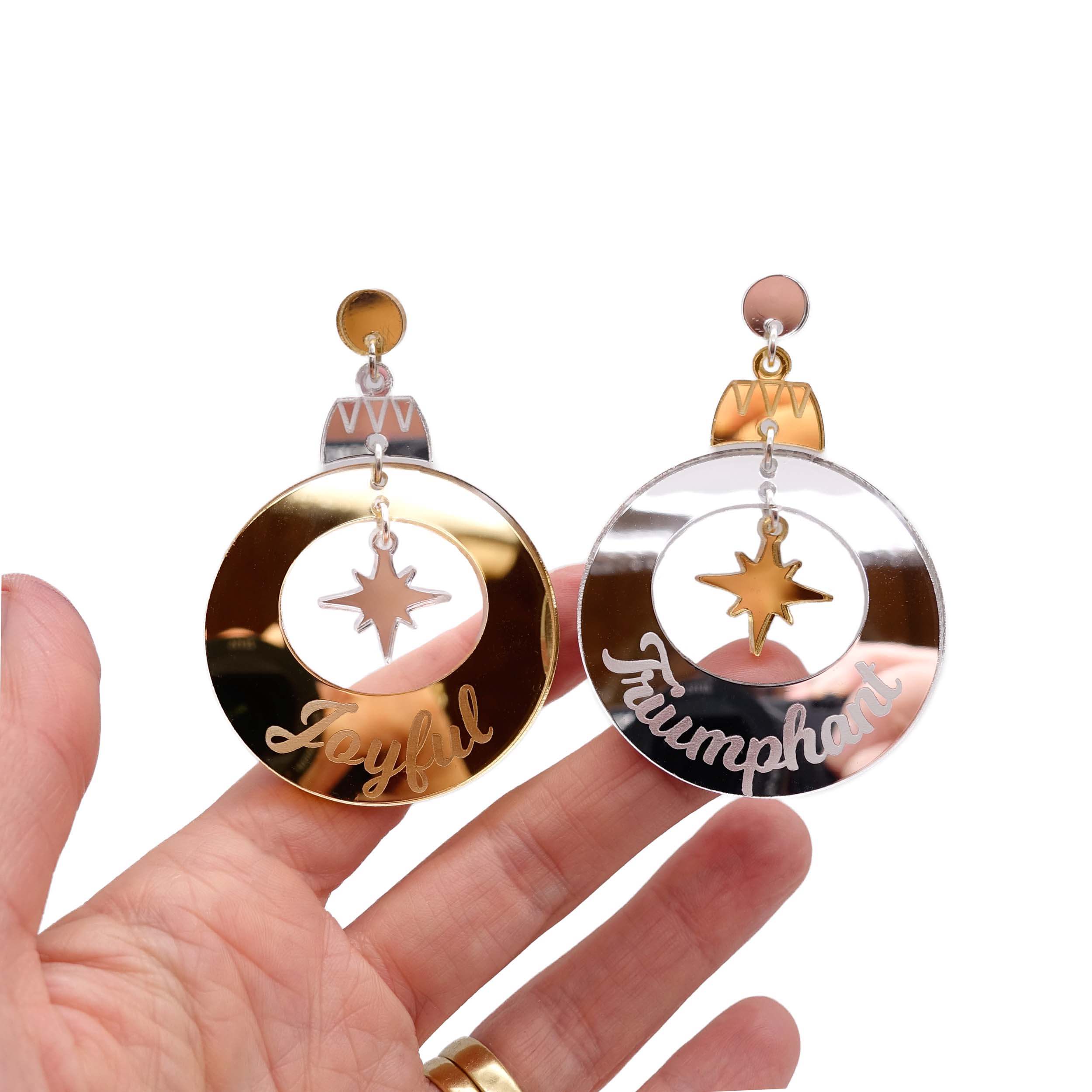 Joyful and Triumphant Christmas Bauble earrings in gold and silver, shown hanging against a white background, with my hand behind for scale and to show the reflective quality of the mirror acrylic.