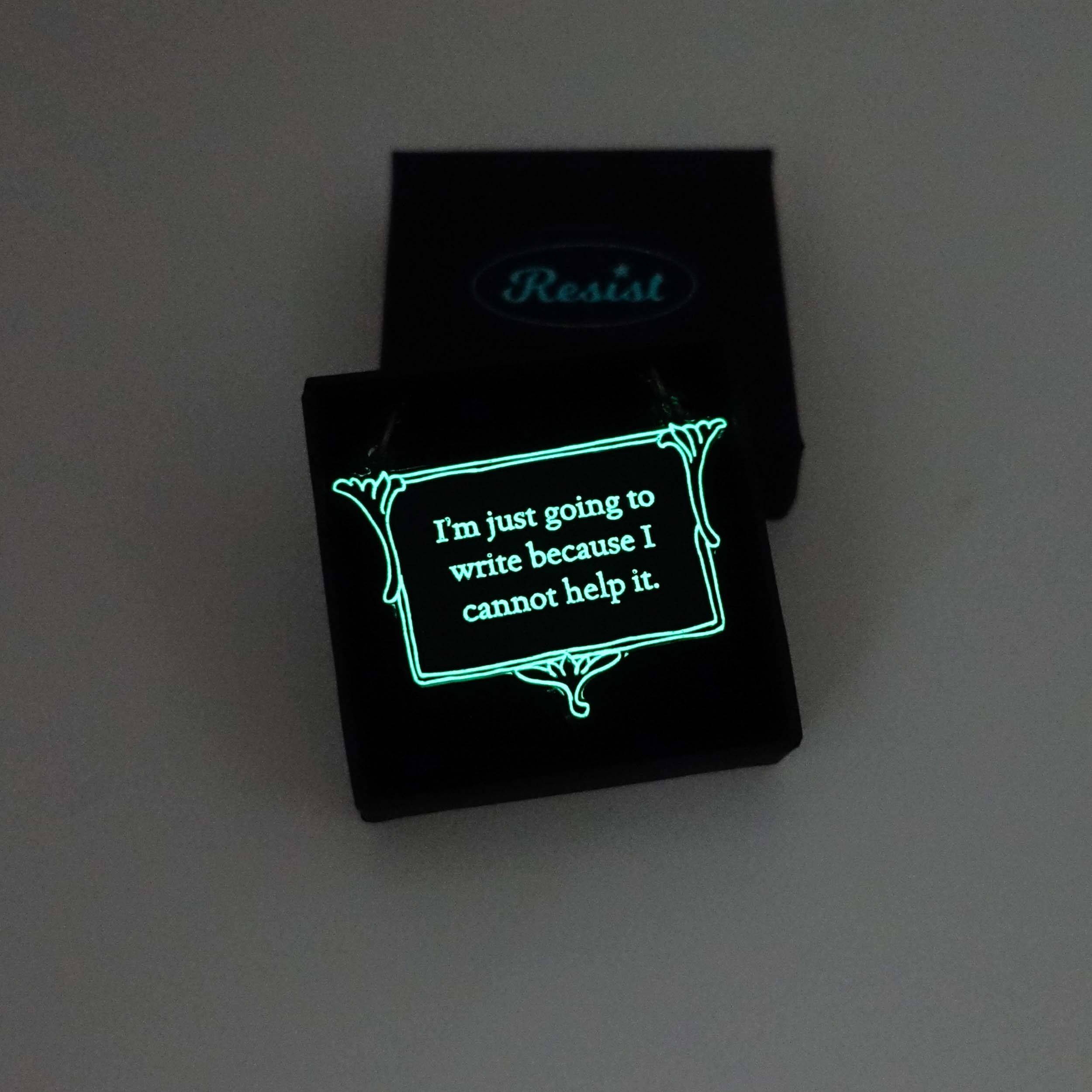 A vintage style slate frost necklace etched with Charlotte Brontë's words in glow-in-the-dark pigment that says, 'I'm just going to write because I cannot help it.' It is shown in a small Wear and Resist gift box glowing in the dark.