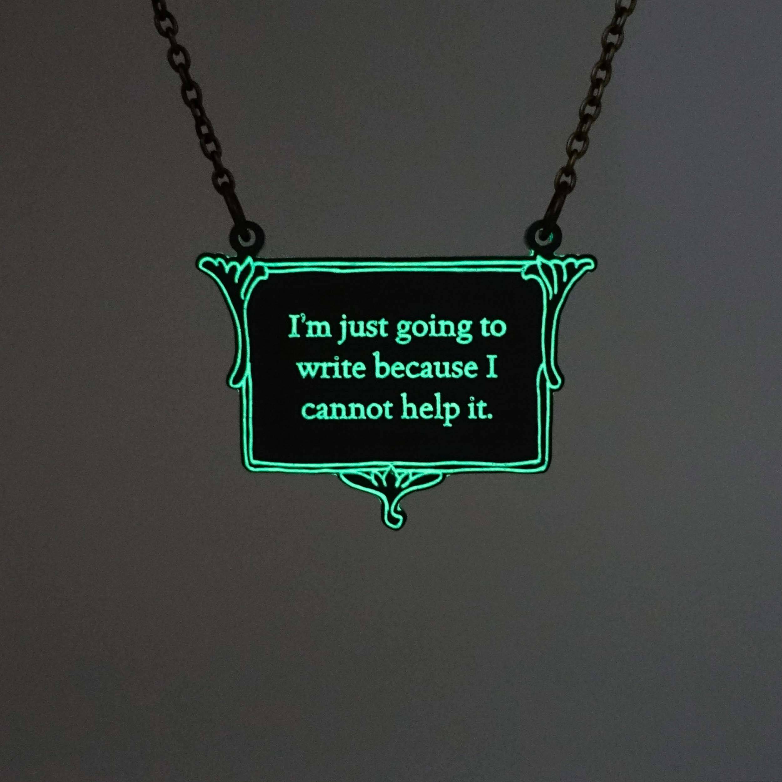A vintage style slate frost necklace etched with Charlotte Brontë's words in glow-in-the-dark pigment that says, 'I'm just going to write because I cannot help it.' It is shown on an antique gold chain glowing in the dark.