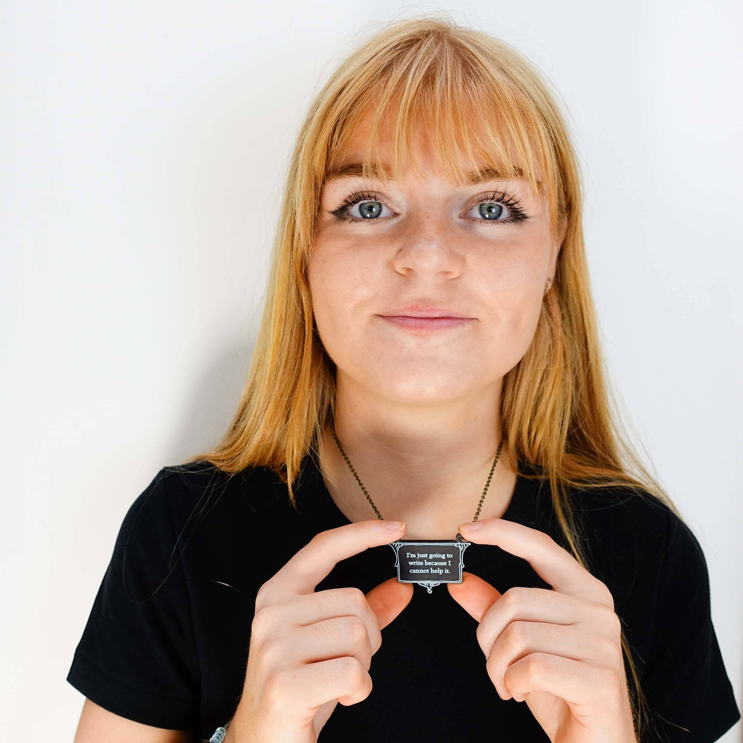 Eliza wears a small vintage style slate frost necklace etched with Charlotte Brontë's words in glow-in-the-dark pigment that says, 'I'm just going to write because I cannot help it.'