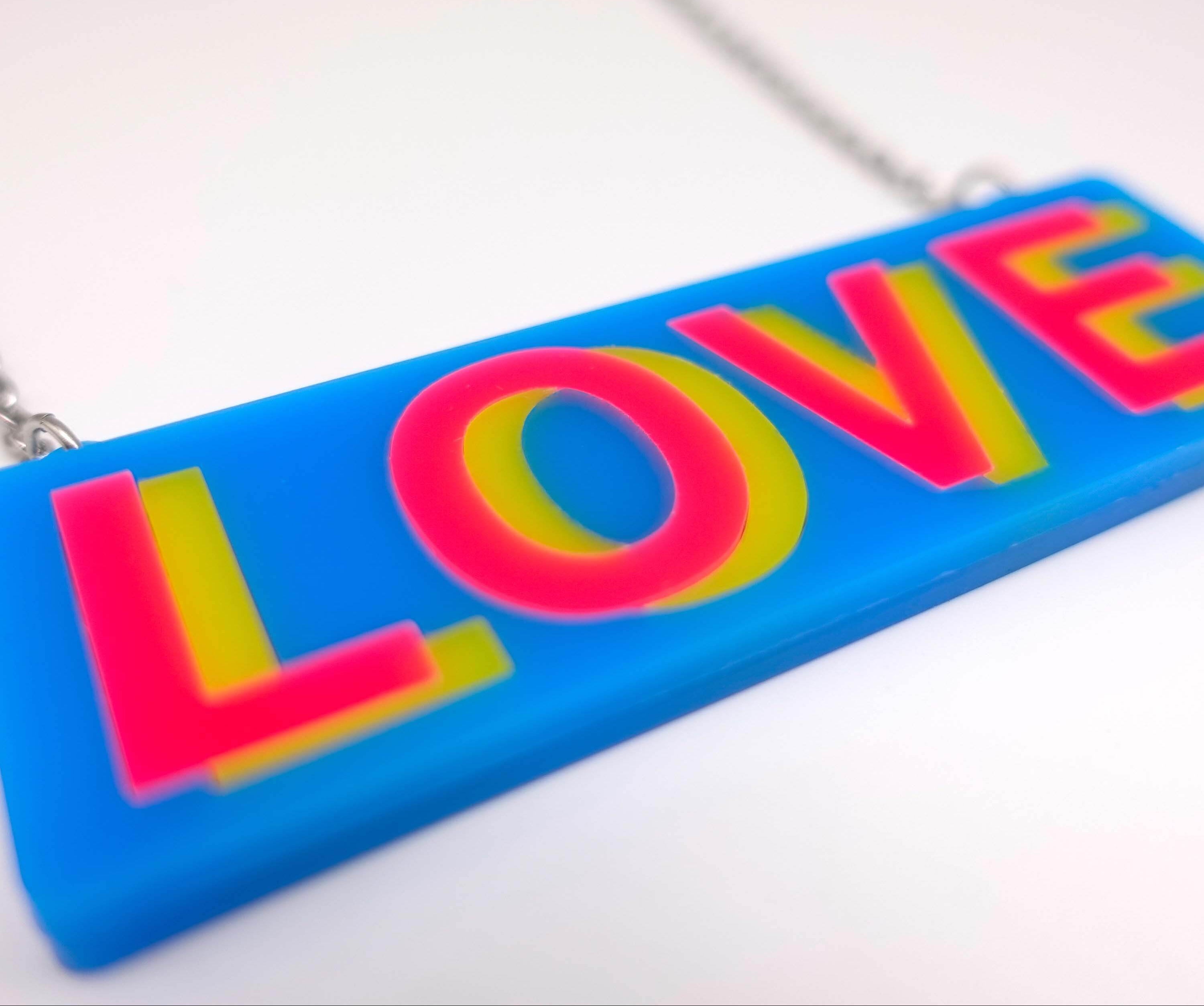 A close up of the LOVE necklace in colourful blue, hot pink and yellow, shown laying on a white surface.