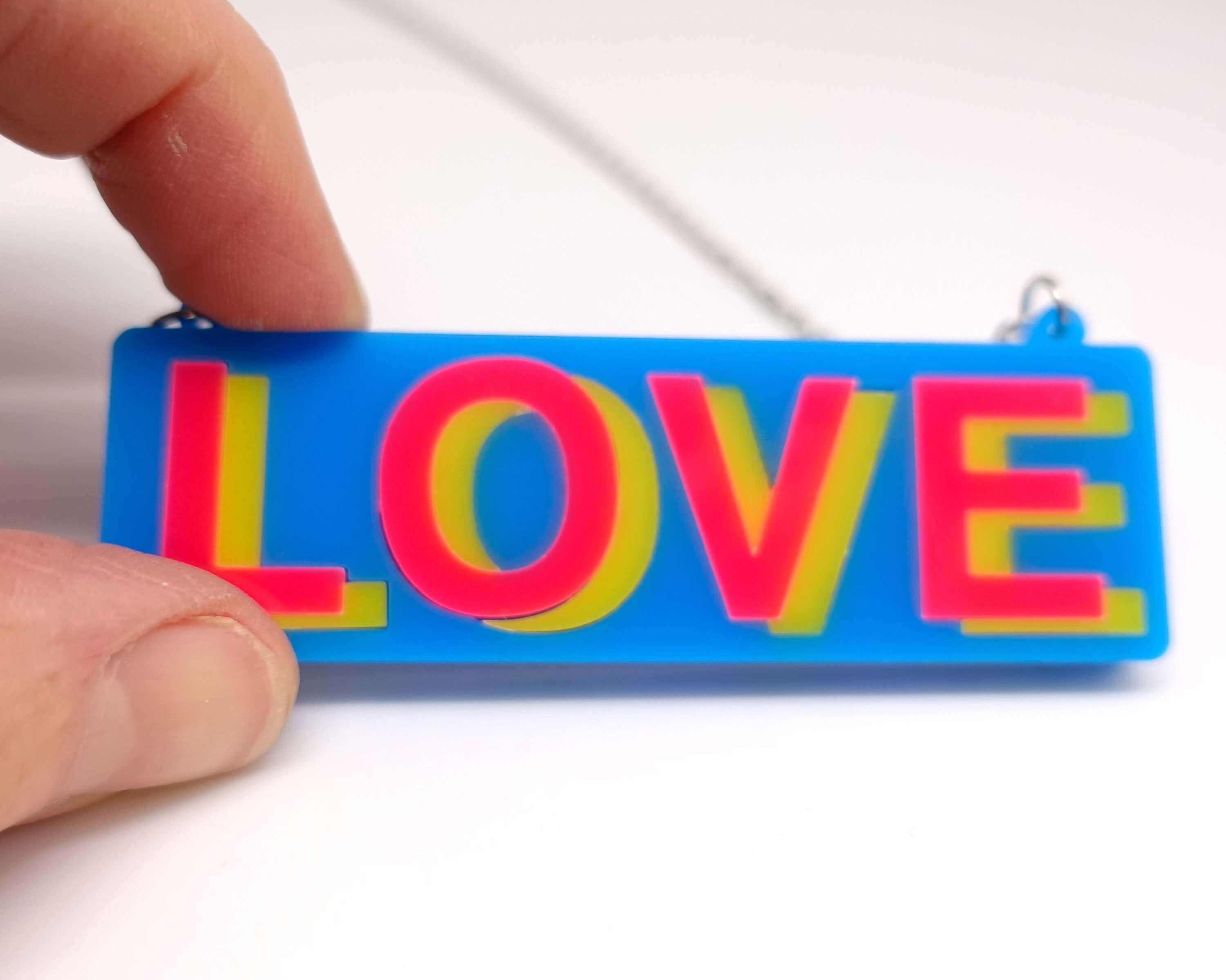 A close up of the LOVE necklace in colourful blue, hot pink and yellow, shown held against a white surface.