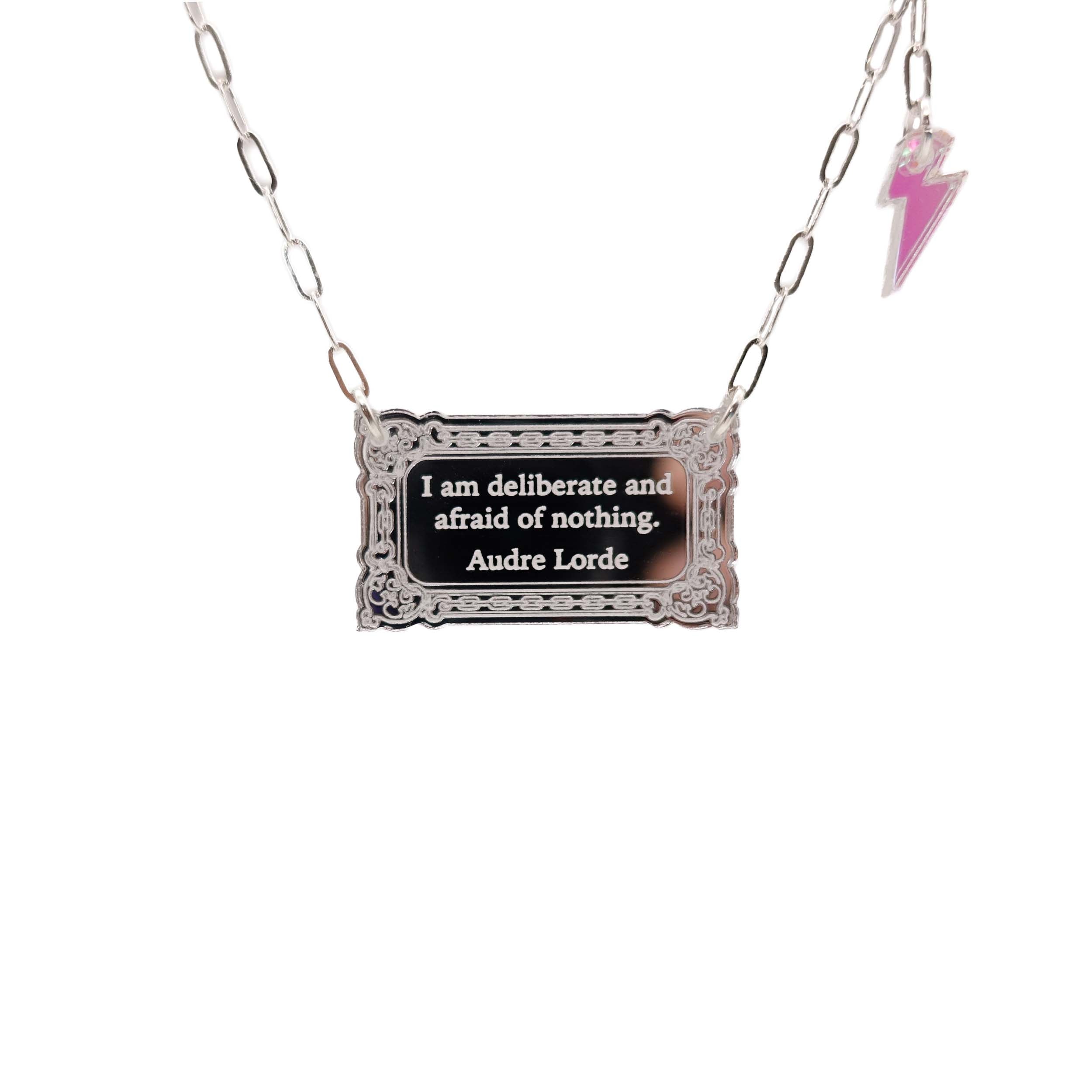 Literary Quote necklace etched with a quote by Audre Lorde, 'I am deliberate and afraid of nothing.' Shown hanging in front of a white background with a small iridescent lightning bolt at the end of the silver paperclip chain.