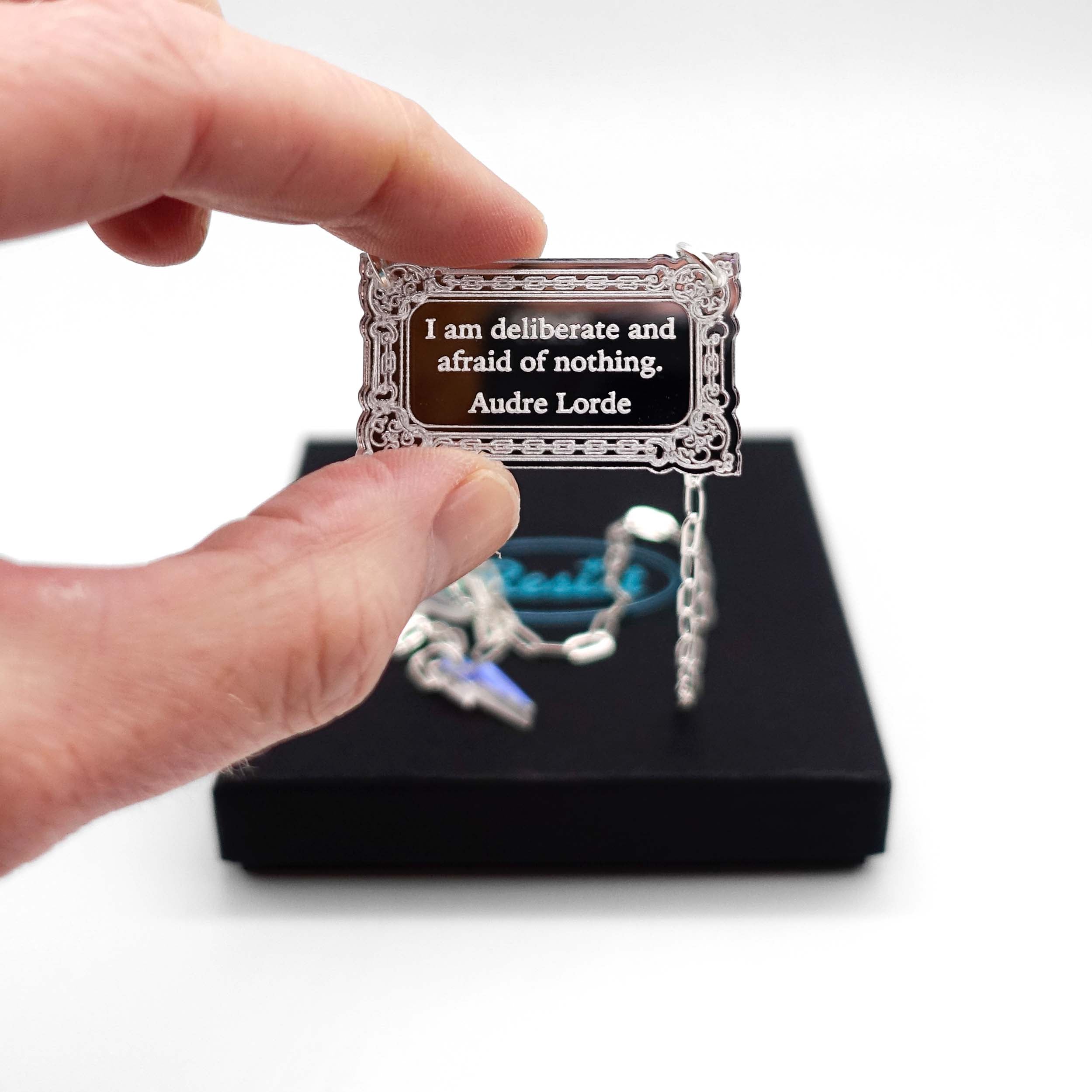 Literary Quote necklace etched with a quote by Audre Lorde, 'I am deliberate and afraid of nothing.' Shown held up in my hand for scale in front of a small Wear and Resist gift box.