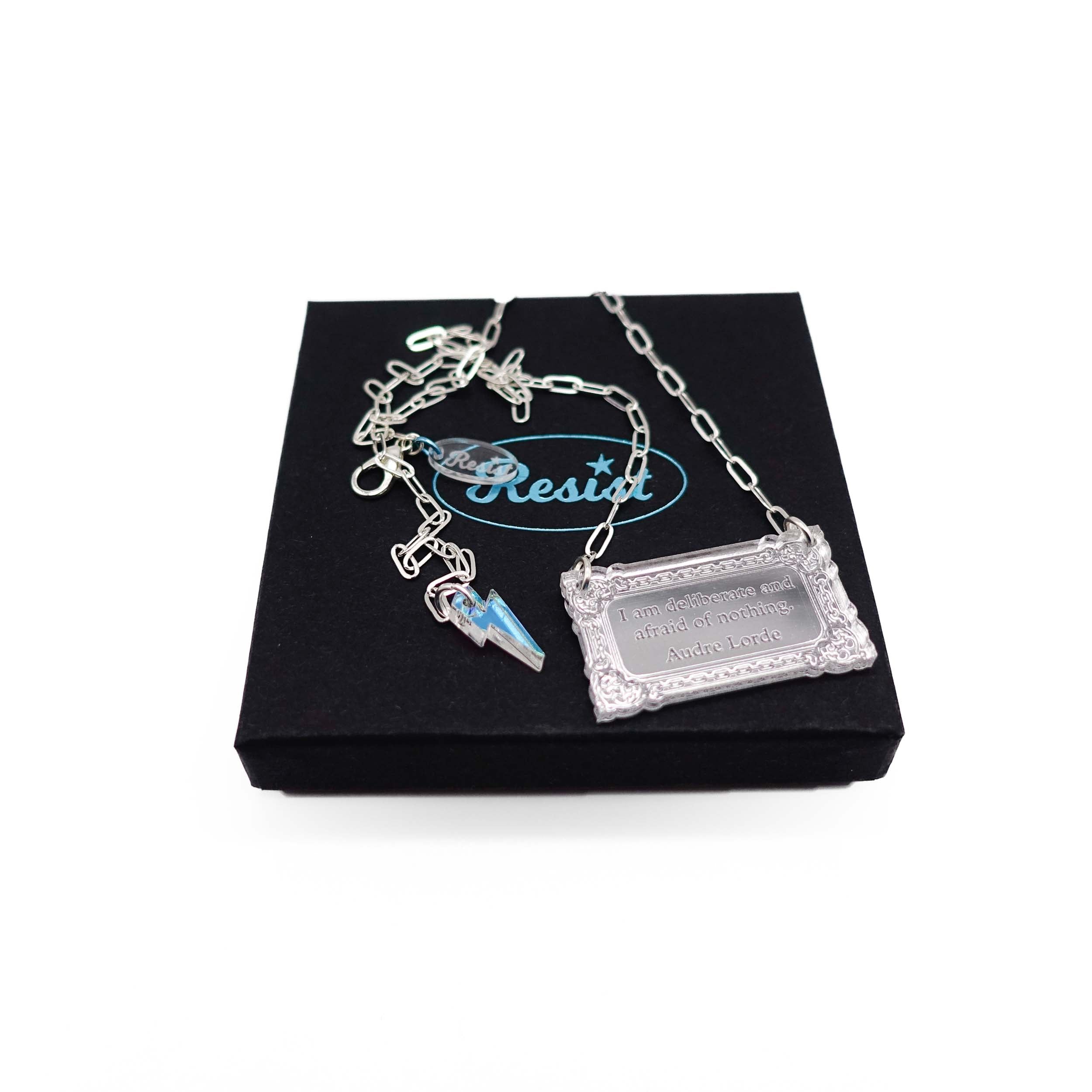 Literary Quote necklace etched with a quote by Audre Lorde, 'I am deliberate and afraid of nothing.' Shown on a silver-plated paperclip chain finished with an iridescent lightning bolt, sitting on a Wear and Resist gift box.
