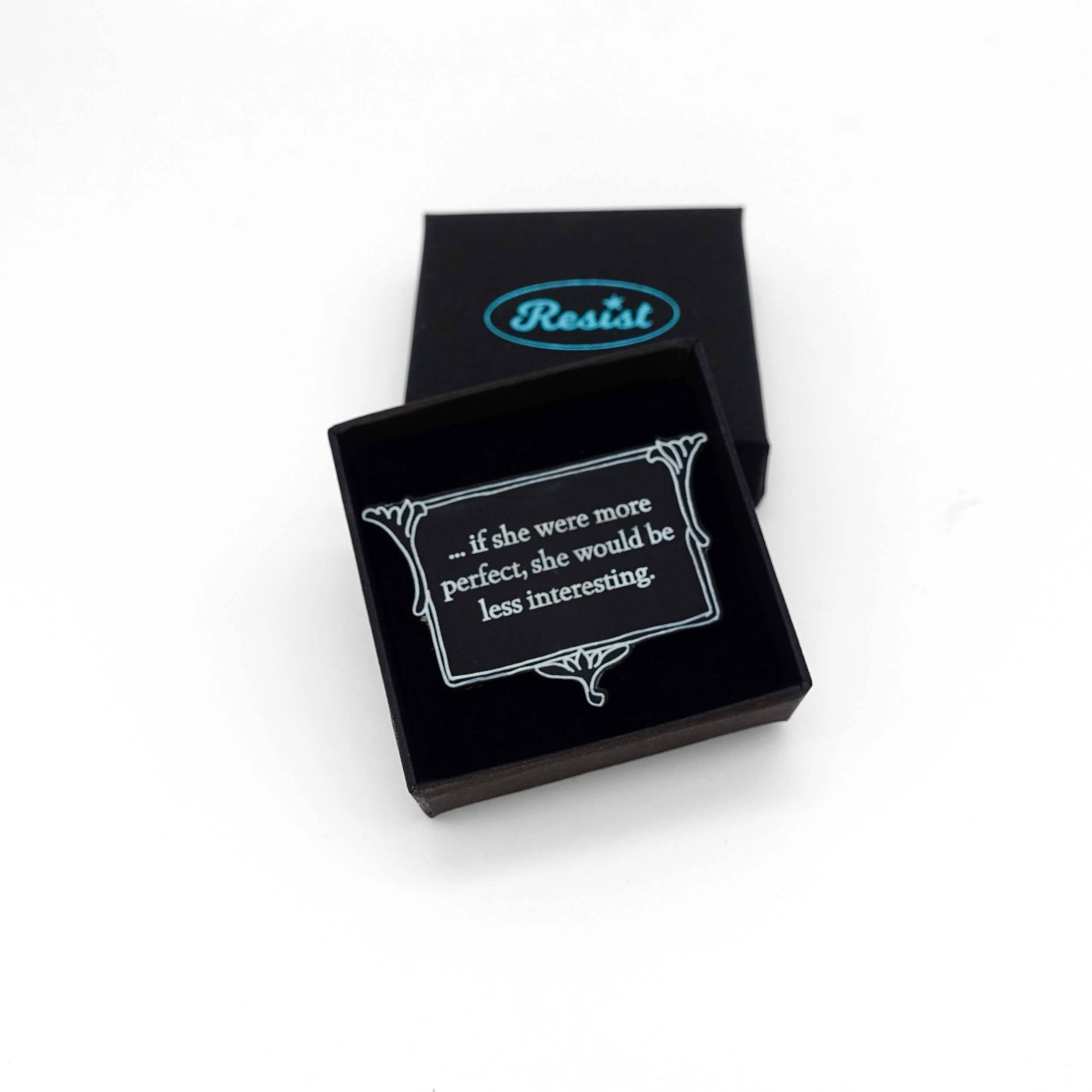 A brooch etched with glow-in-the-dark pigment that says, 'if she were more perfect, she would be less interesting' from Anne Brontë's The Tenant of Wildfell Hall. It is shown in a small Wear and Resist gift box.