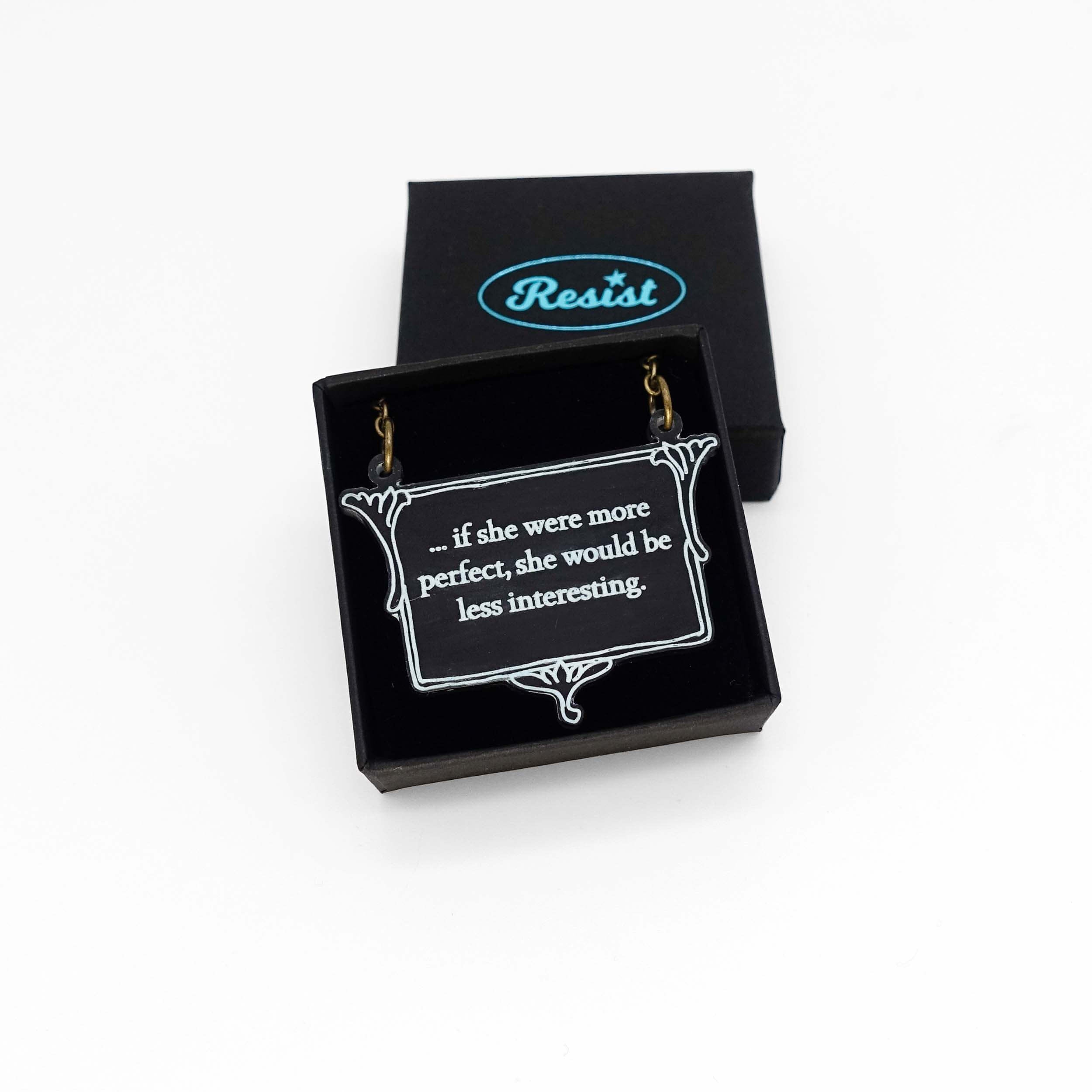 A vintage style slate frost necklace etched with glow-in-the-dark pigment that says, 'if she were more perfect, she would be less interesting' from Anne Brontë's The Tenant of Wildfell Hall, shown in a small Wear and Resist gift box.
