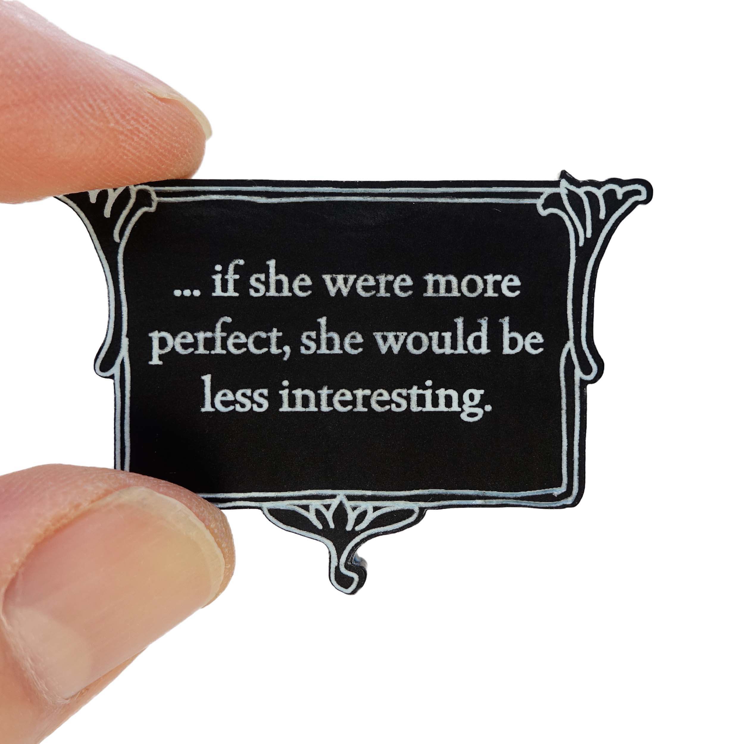 A matte black vintage-style brooch etched with glow-in-the-dark pigment that says, 'if she were more perfect, she would be less interesting' from Anne Brontë's The Tenant of Wildfell Hall, shown held up in my fingers for scale.