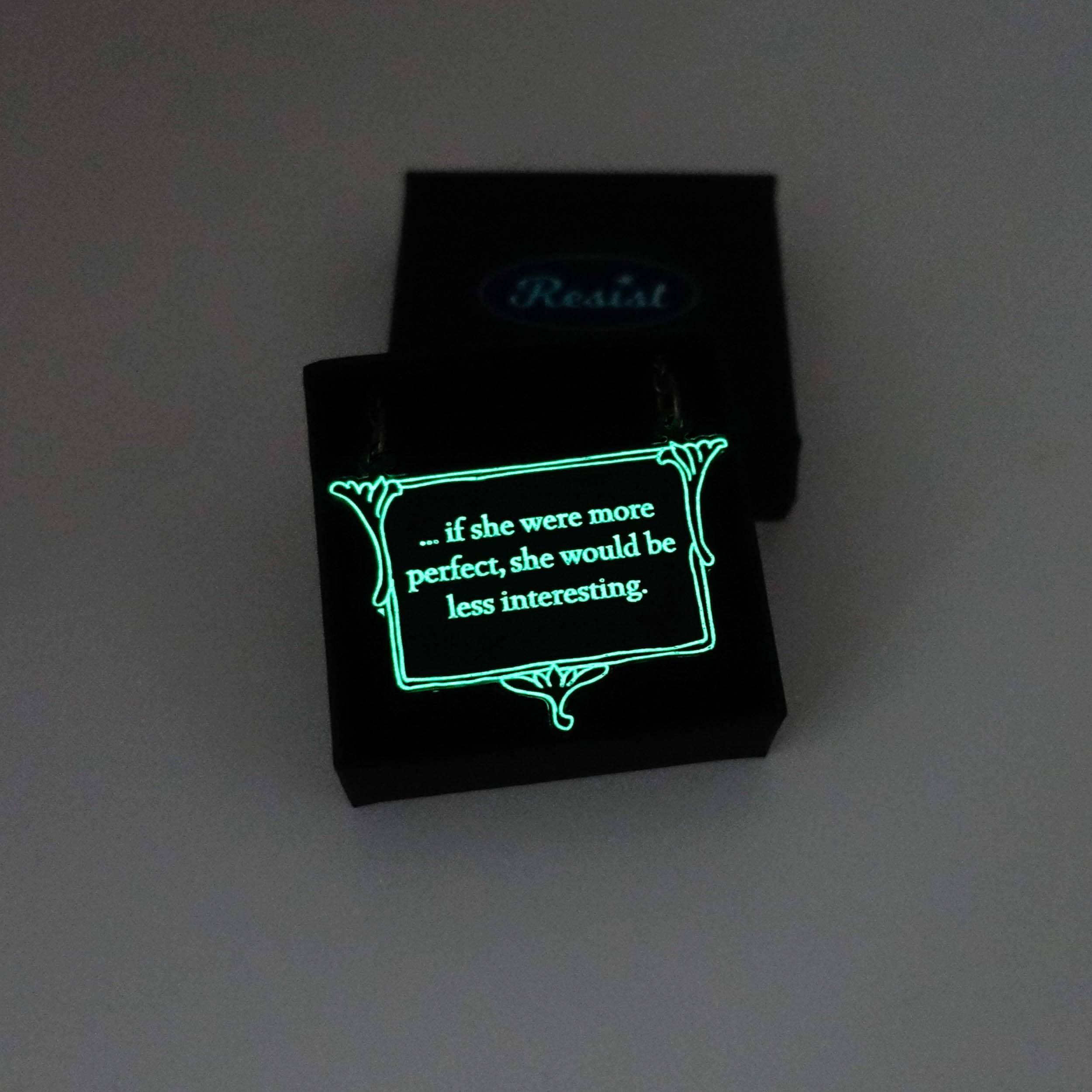 A vintage style slate frost necklace etched with glow-in-the-dark pigment that says, 'if she were more perfect, she would be less interesting' from Anne Brontë's The Tenant of Wildfell Hall, shown glowing in the dark in a Wear and Resist gift box.