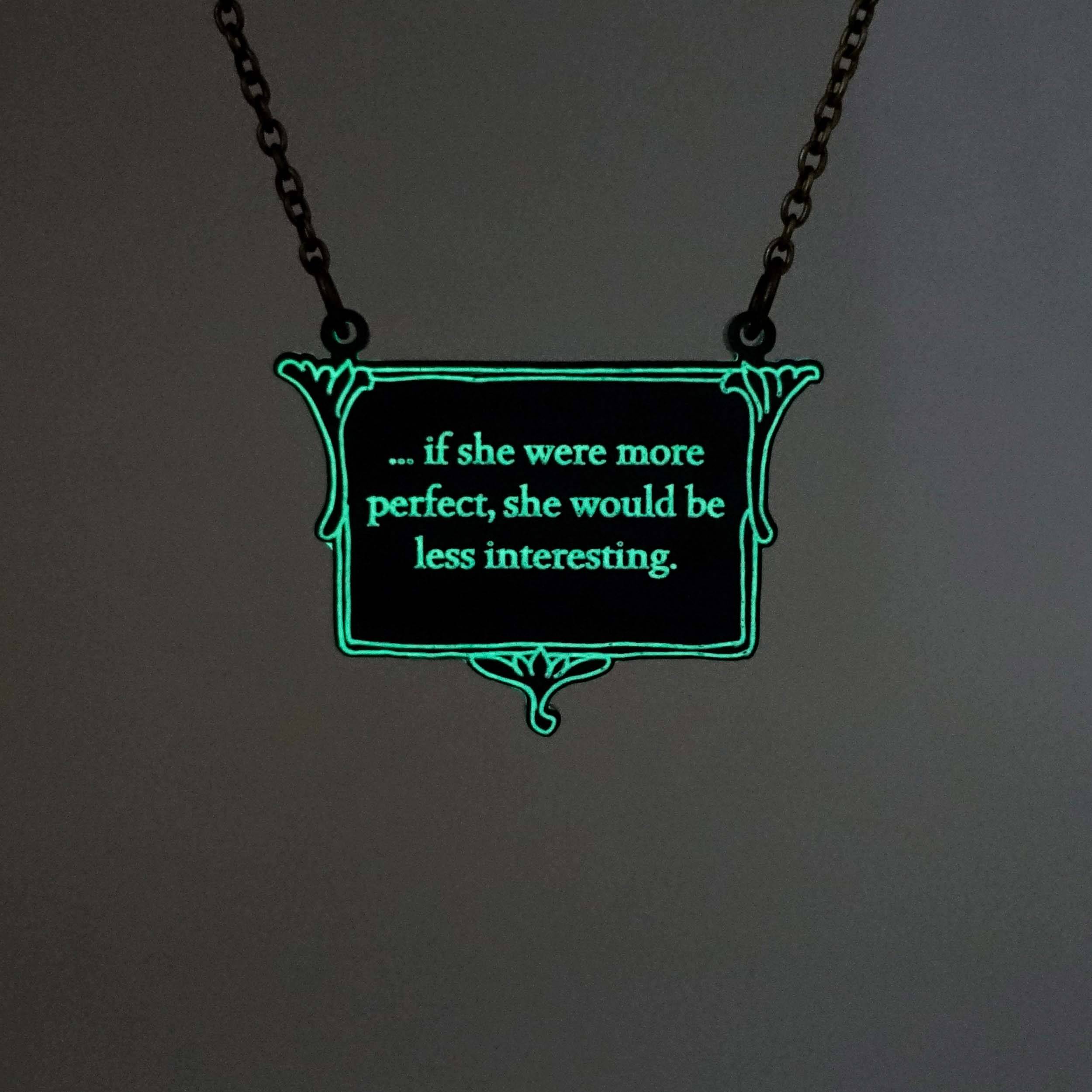 A vintage style slate frost necklace etched with glow-in-the-dark pigment that says, 'if she were more perfect, she would be less interesting' from Anne Brontë's The Tenant of Wildfell Hall, shown on an antique gold chain glowing in the dark.