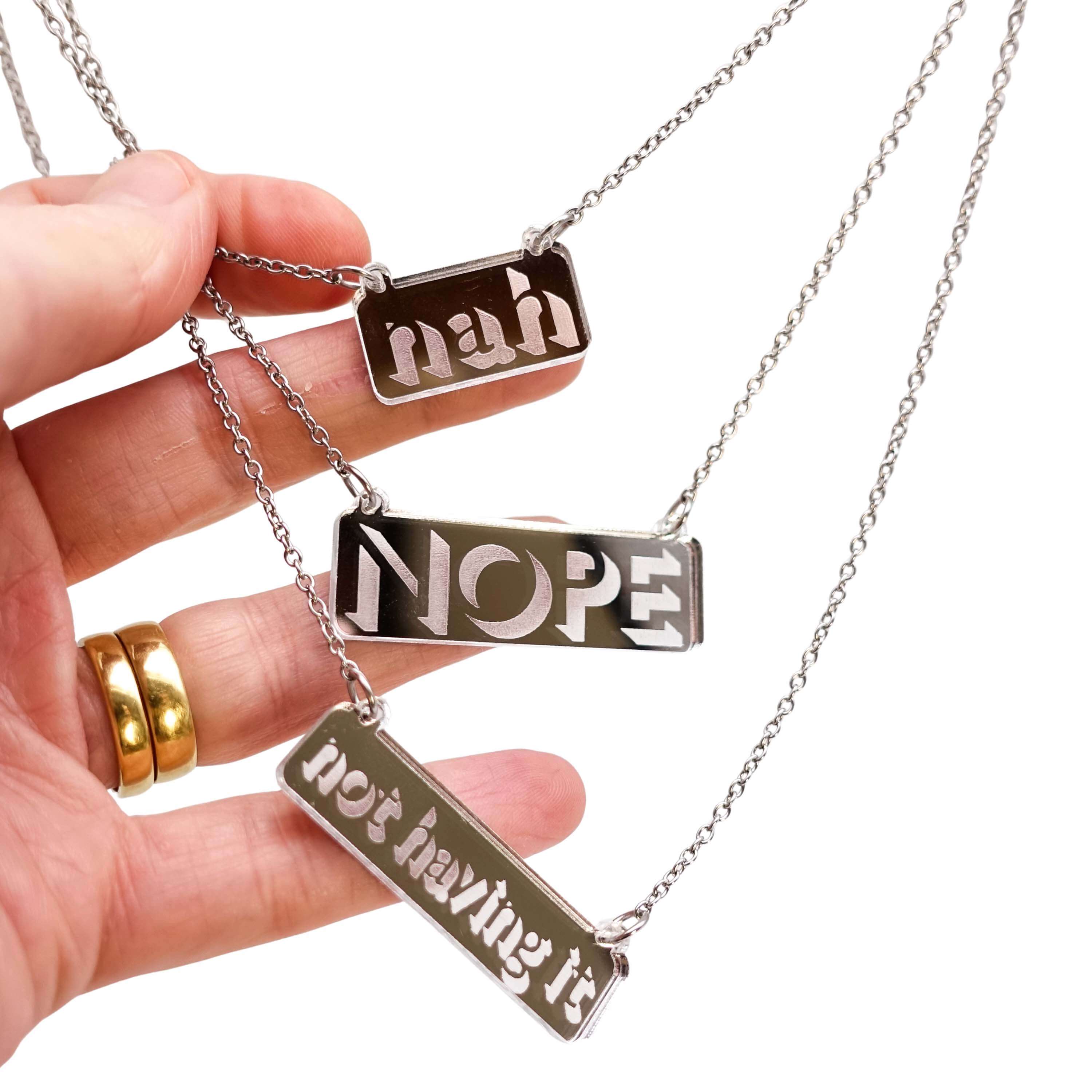 Three necklaces in etched silver mirror that say Nah, Nope and Not Having It, hanging on stainless steel chains with my hand holding them for scale.