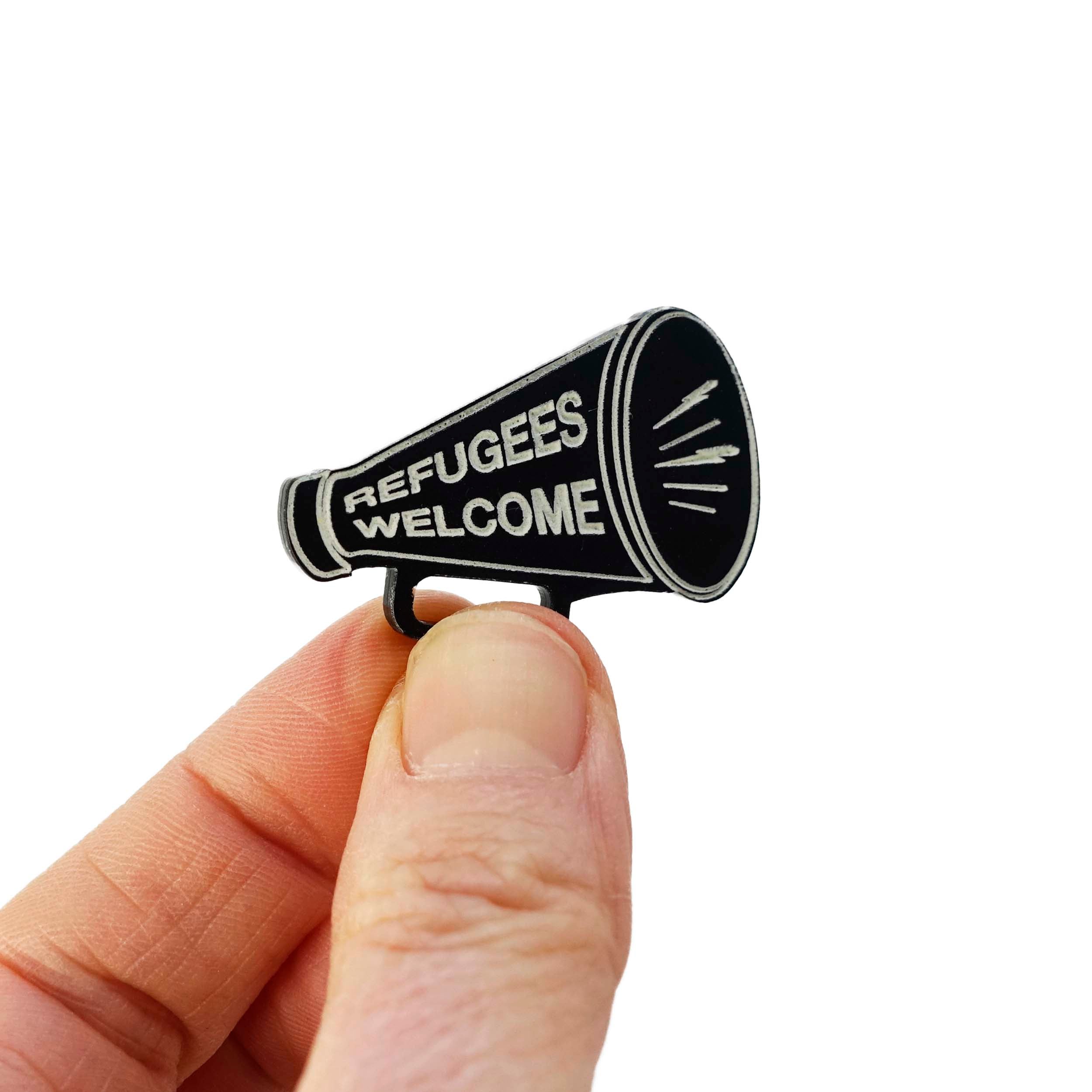 Small megaphone brooch in matte black etched with light glow-in-the-dark paint with the words Refugees Welcome. Shown held up in my fingers for scale.