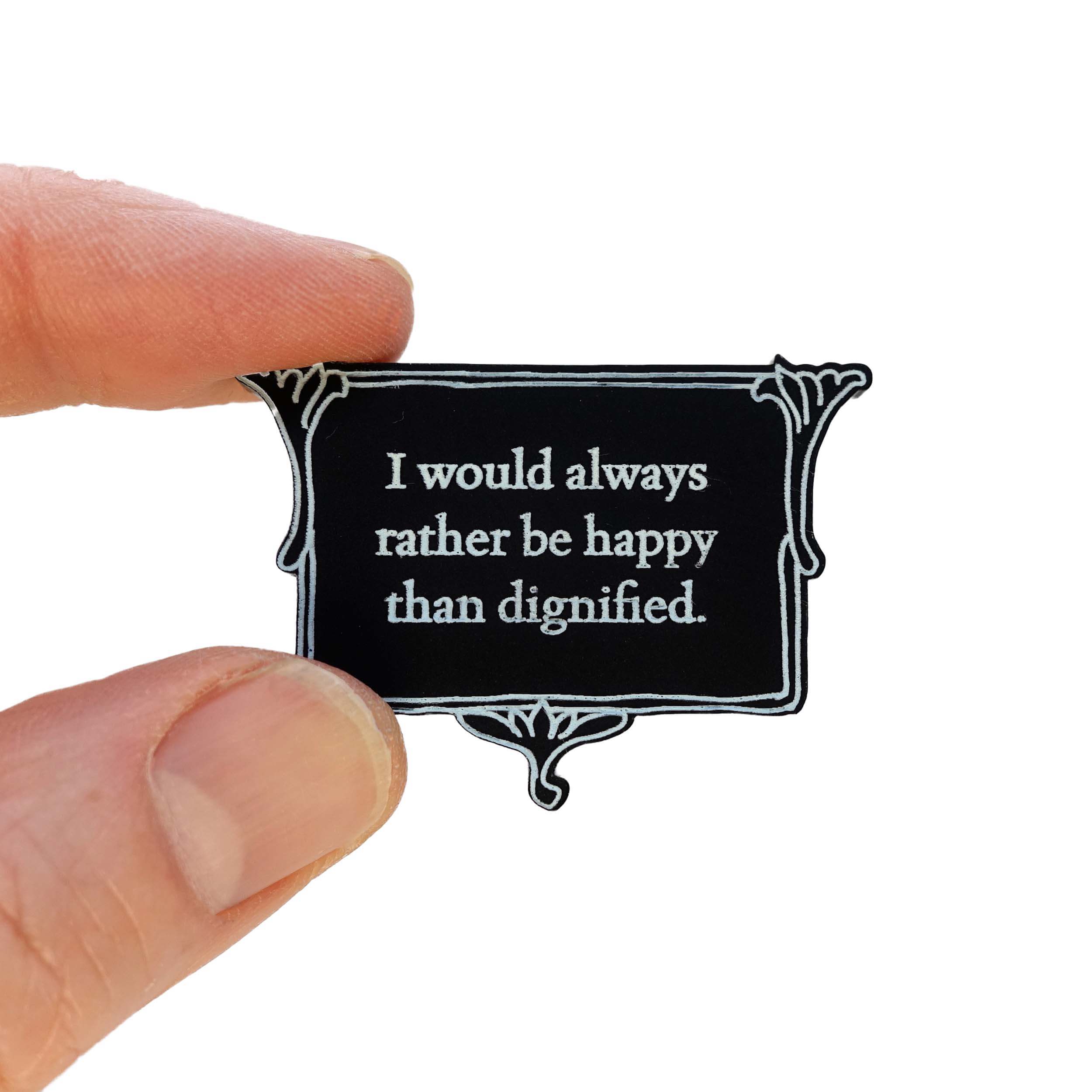 A matte black brooch etched with glow-in-the-dark pigment that says, 'I would always rather be happy than dignified.'  A quote from Charlotte Brontë's Jane Eyre from The Brontë Collection designed by Wear and Resist, shown held up in my fingers for scale.