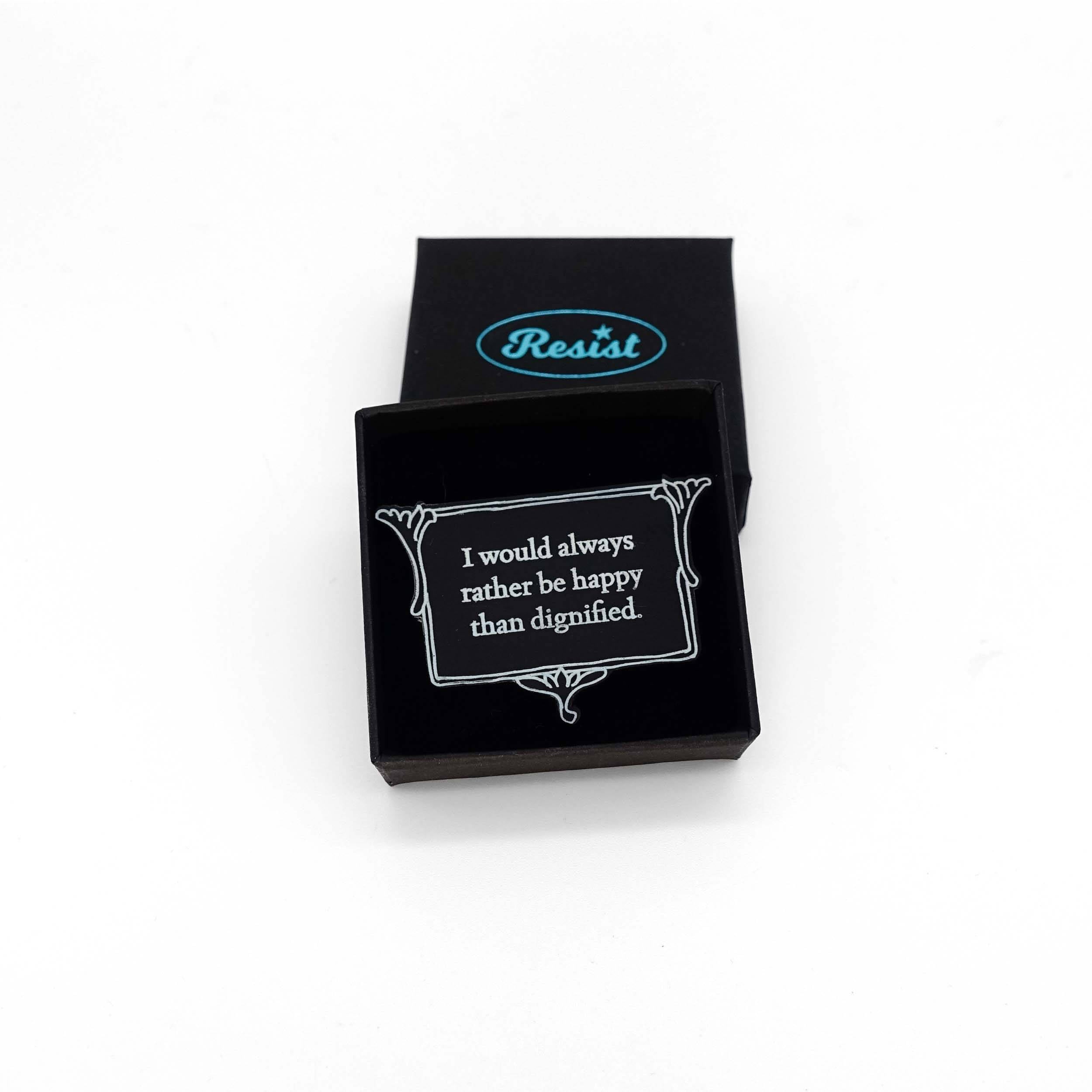 Matte black brooch etched with glow-in-the-dark pigment that says, 'I would always rather be happy than dignified.'  A quote from Charlotte Brontë's Jane Eyre from The Brontë Collection designed by Wear and Resist, shown in a small Wear and Resist gift box.