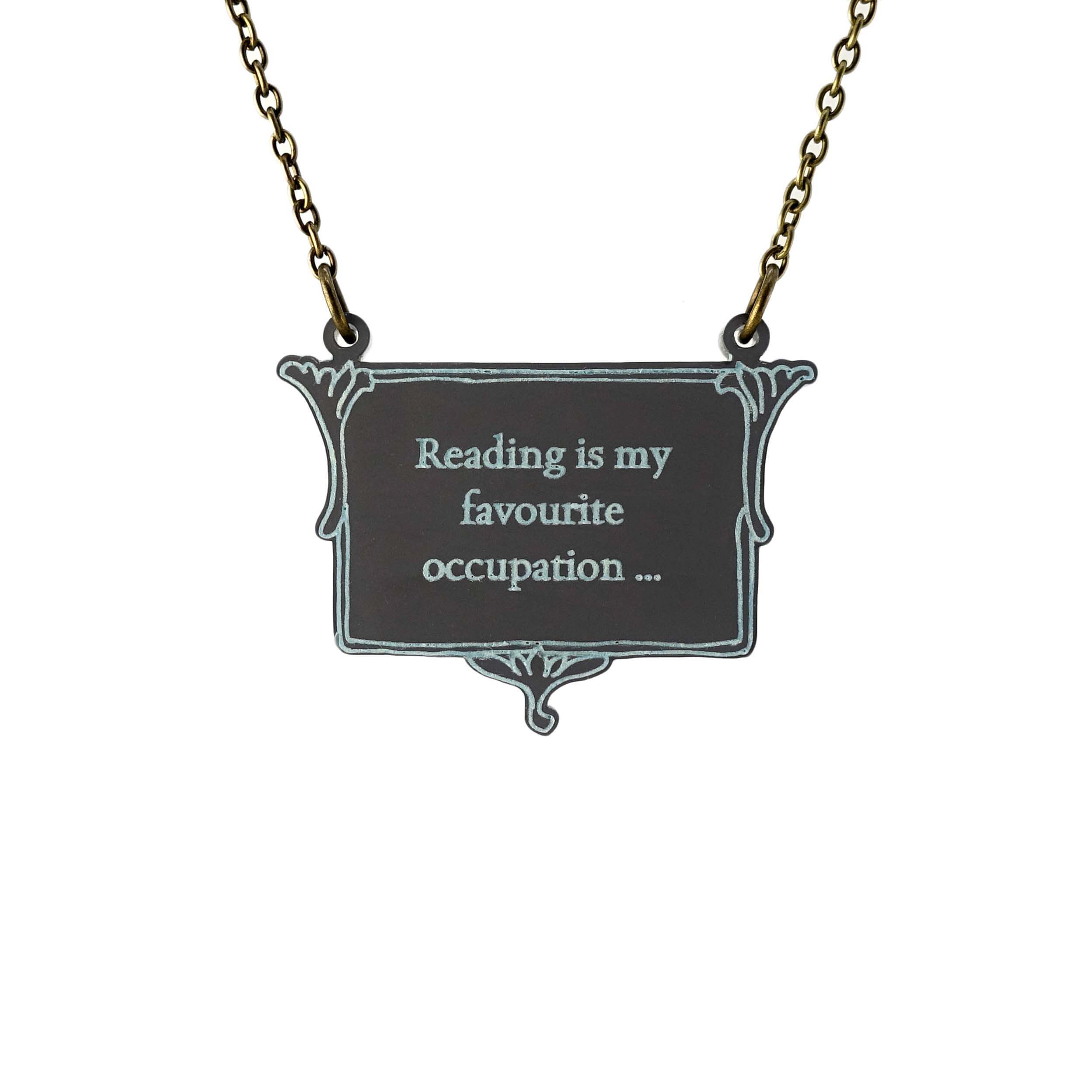 A vintage style slate frost necklace etched with glow-in-the-dark pigment that says, 'Reading is my favourite occupation ...' a quote from Anne Brontë's Agnes Grey, shown on an antique gold chain hanging against a white backround.