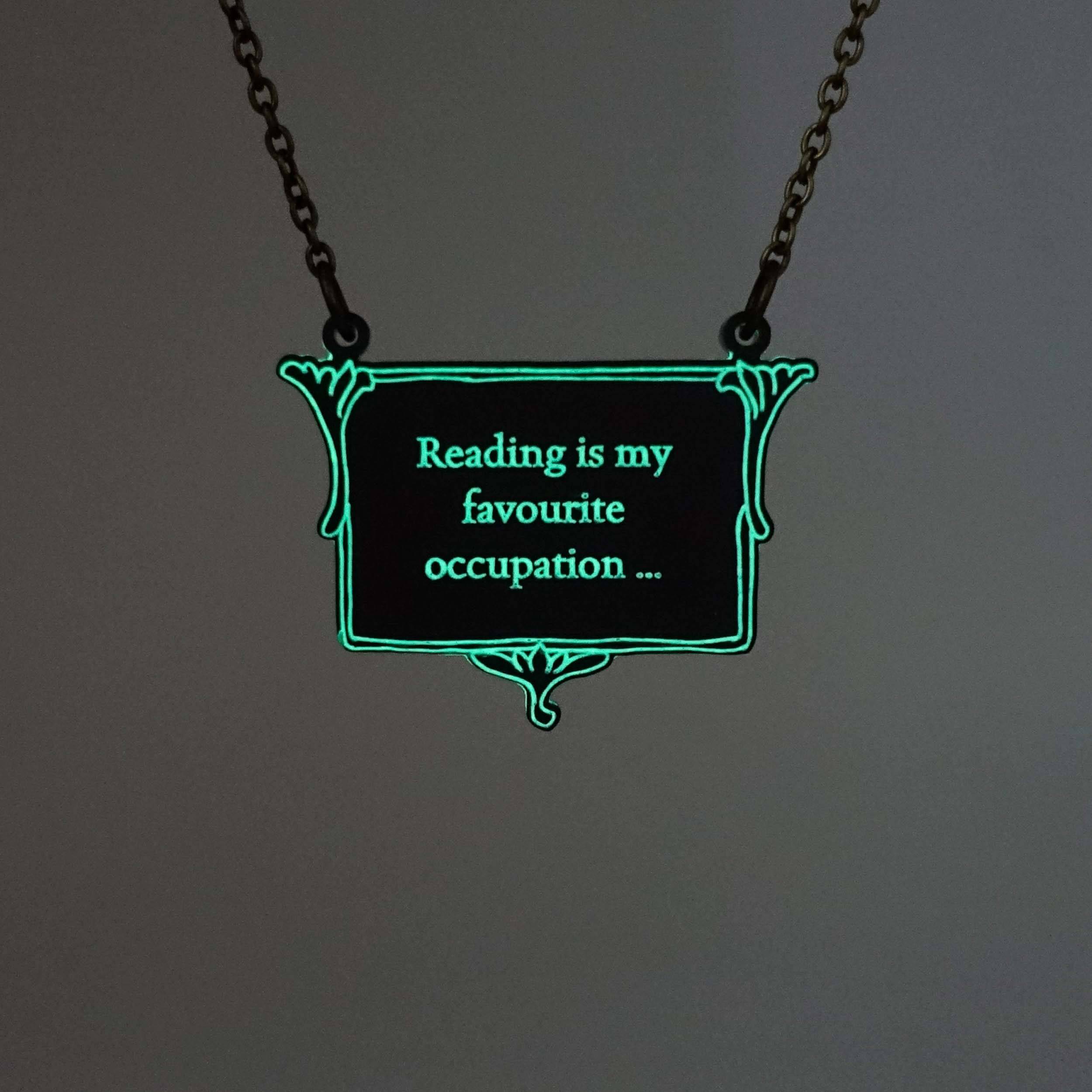 A vintage style slate frost necklace etched with glow-in-the-dark pigment that says, 'Reading is my favourite occupation ...' a quote from Anne Brontë's Agnes Grey, shown on an antique gold chain glowing in the dark.