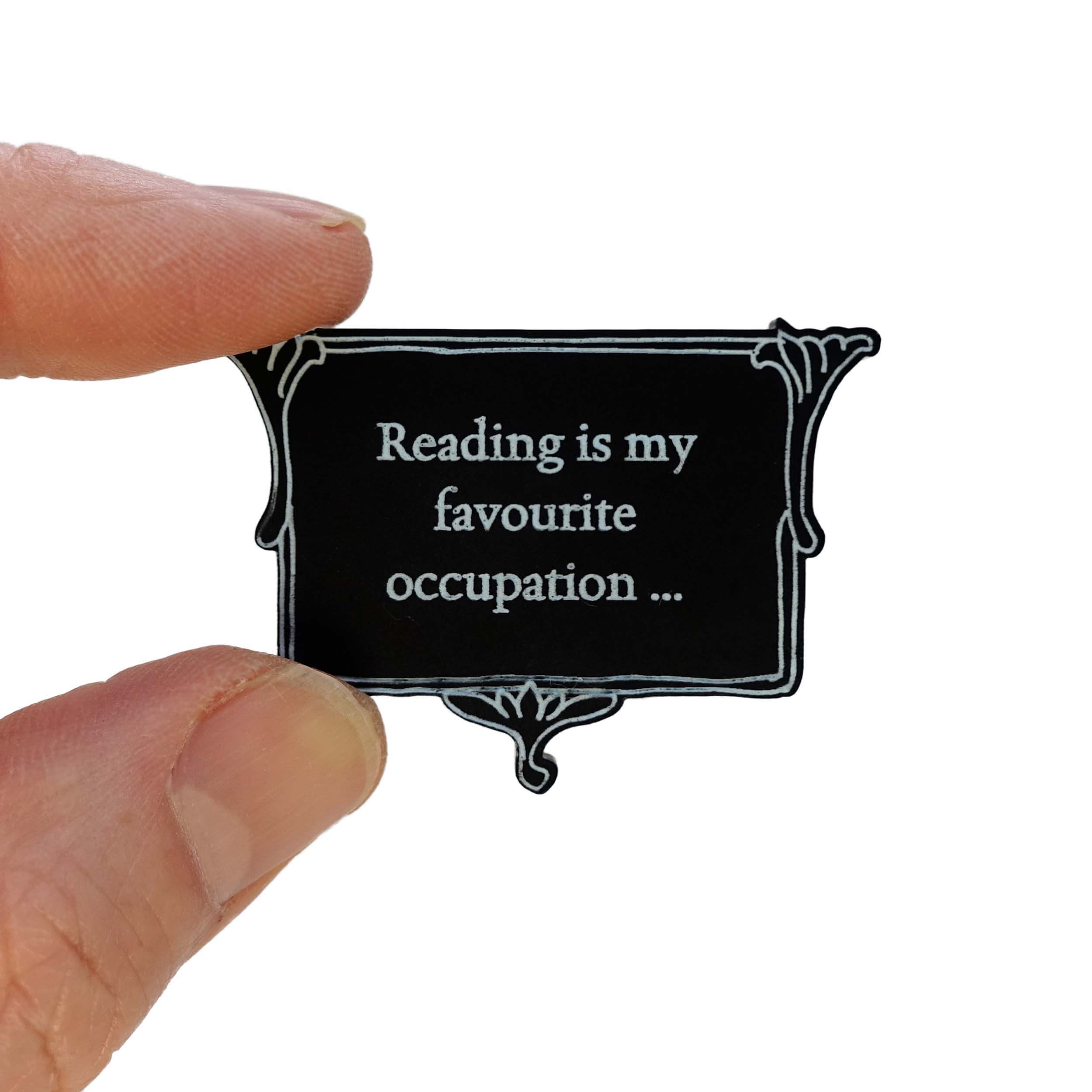 A matte black brooch etched with glow-in-the-dark pigment that says,  'Reading is my favourite occupation ...' a quote from Anne Brontë's Agnes Grey, held up in my fingers for scale.