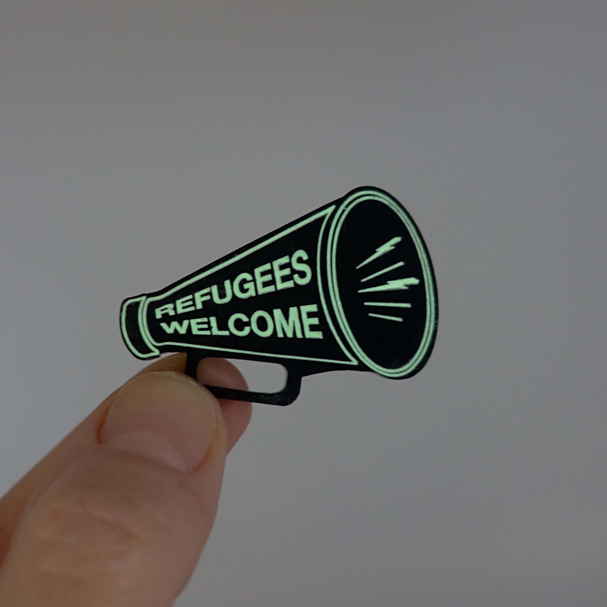 Small megaphone brooch in matte black etched with light glow-in-the-dark paint with the words Refugees Welcome, glowing in the dark. Shown held up in my fingers for scale.