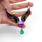 My hand holding the Resist Persist winged heart necklace so the light catches it.