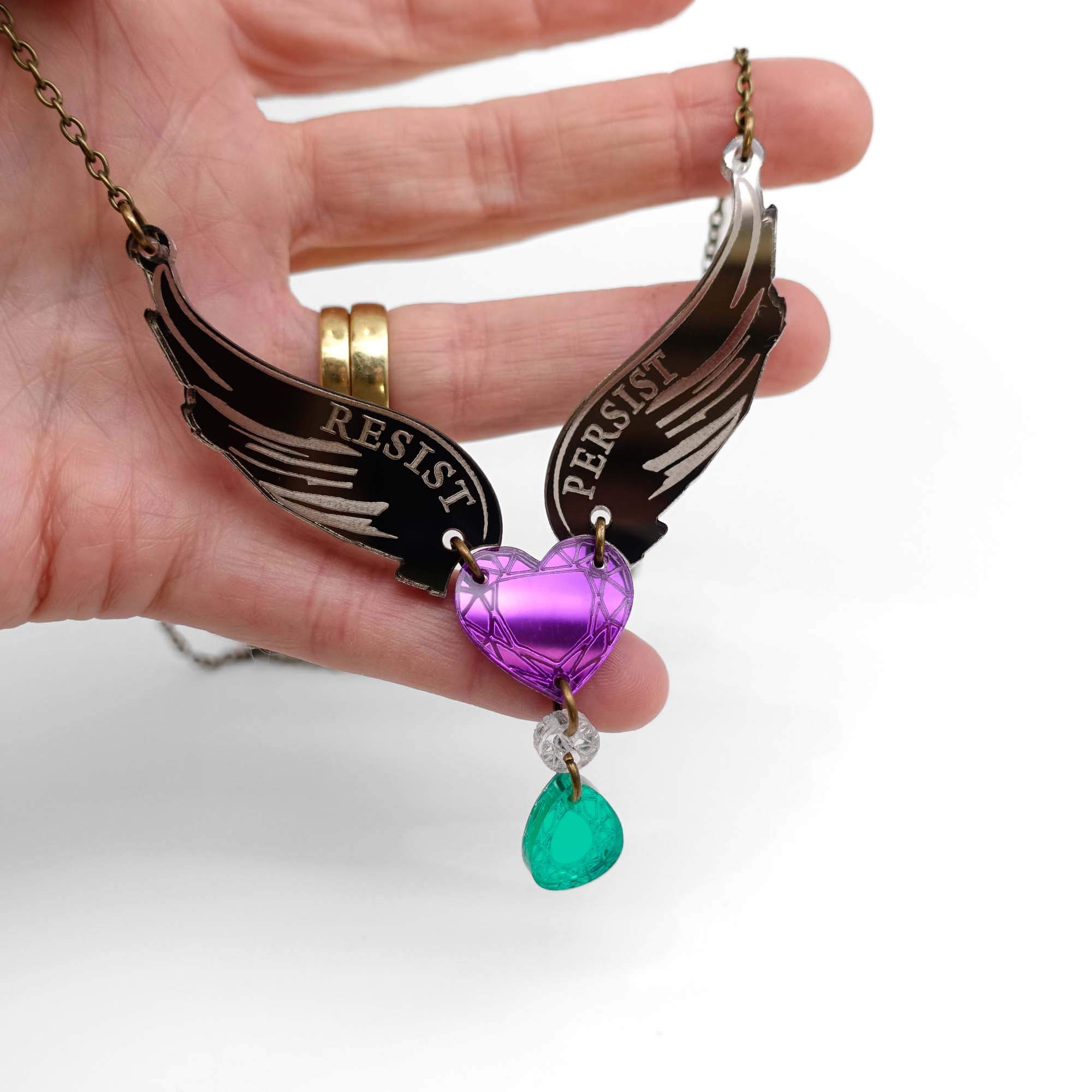 My hand holding the Resist Persist winged heart necklace so the light catches it.