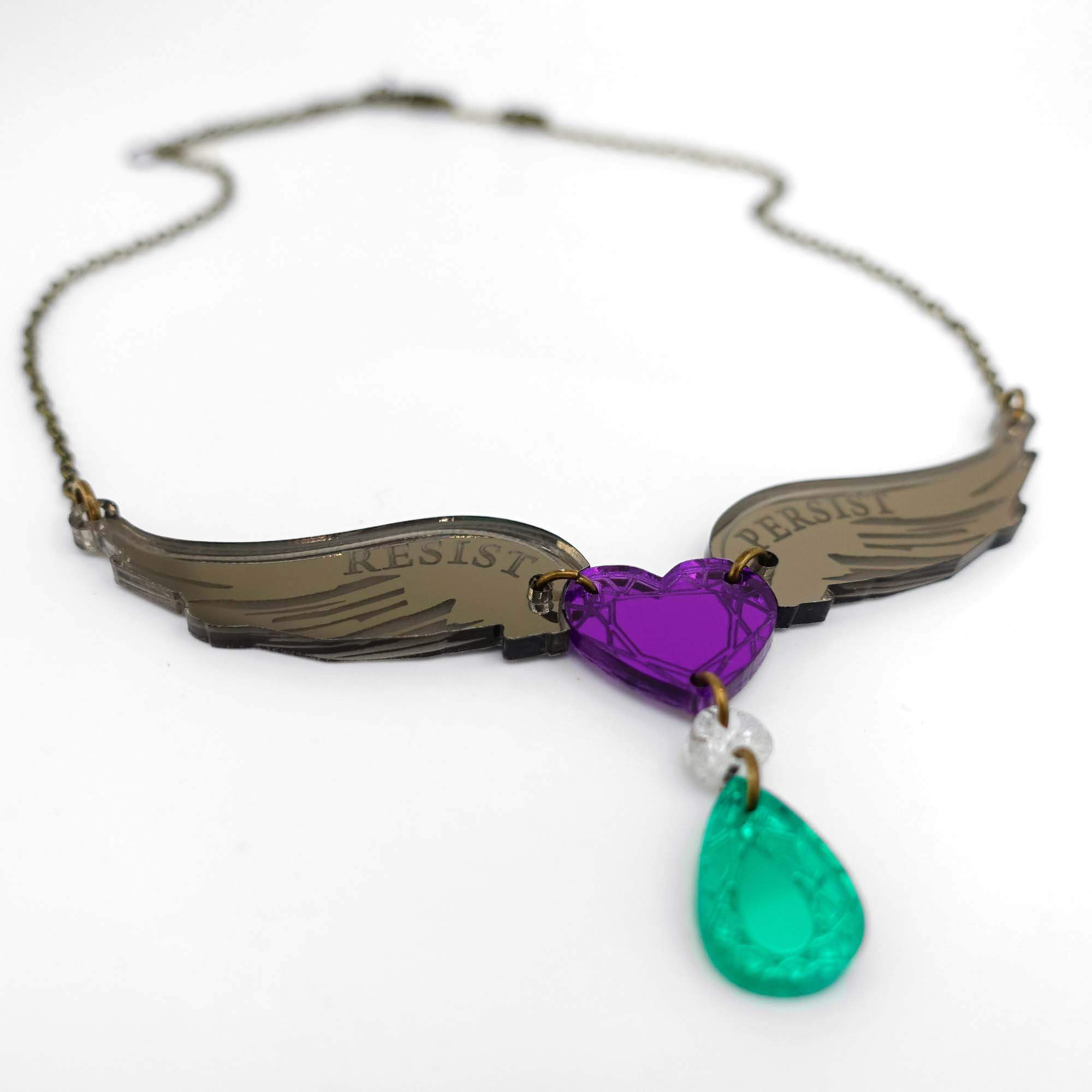 A necklace with bronze wings around a purple heart etched with the words, Resist Persist, and a white and green acrylic jewell hanging from the heart in the Suffragette colours. Shown lying on a white surface.